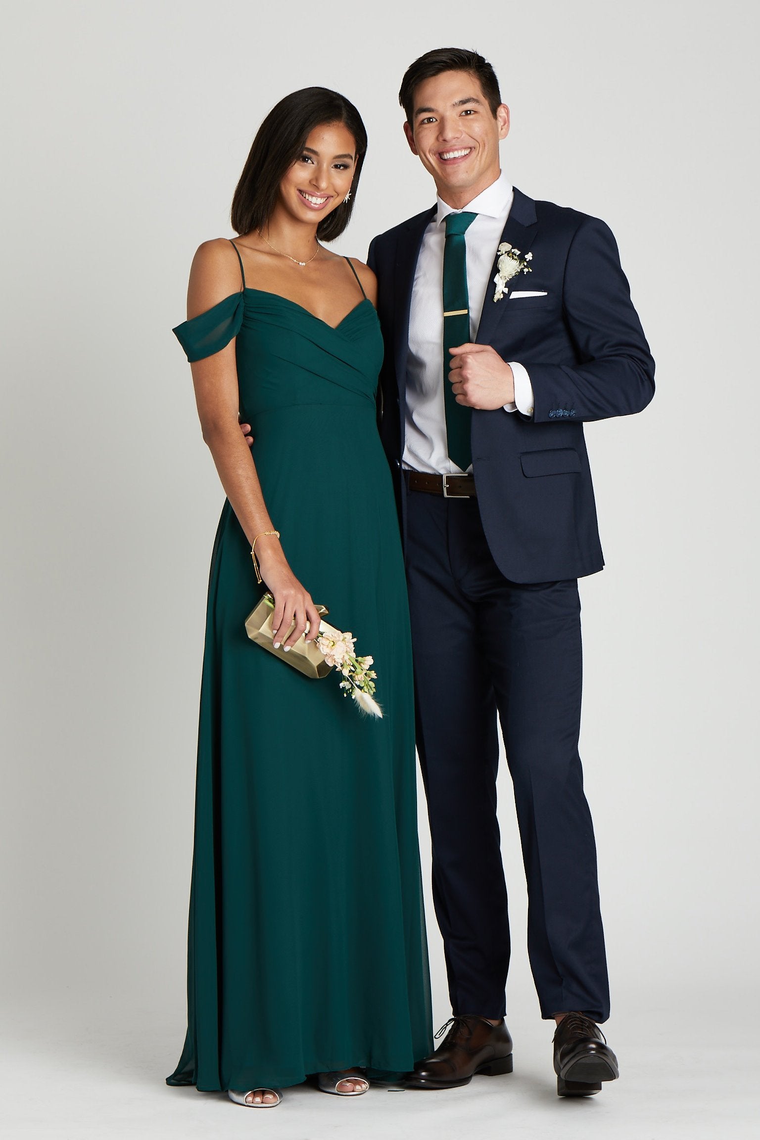 Front view  of two models in a side embrace at the waist. One model with a slender physique and a medium skin tone wears the Simon Necktie in emerald with a white collared button down shirt and the Navy Blue Suit by Suitshop. The second model with a slender figure and a medium skin tone coordinates their look wearing a floor length dress in emerald.