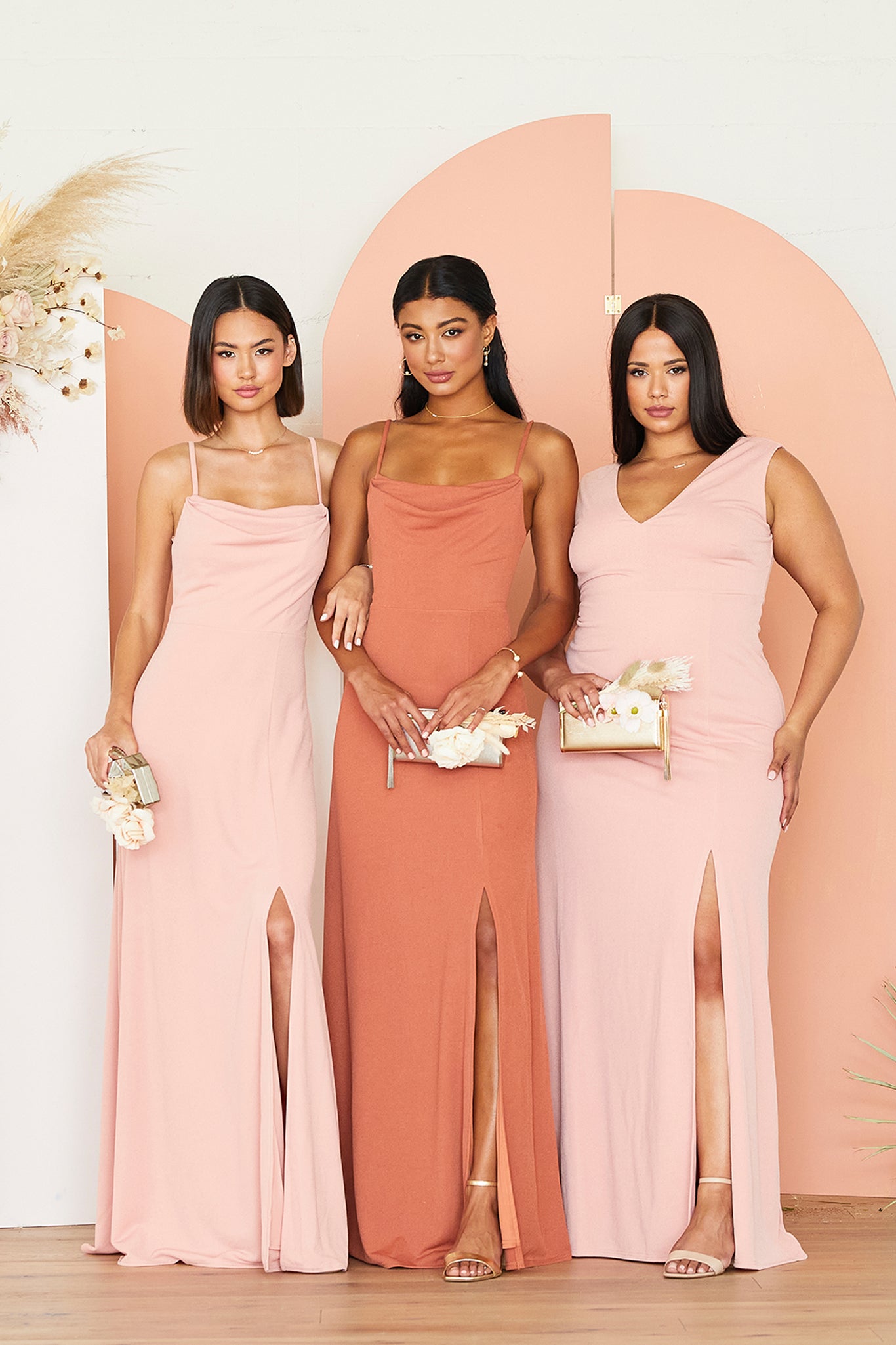 Front view of three models standing together and wearing bridesmaid dresses in shades of pink. A slender model with a medium-light skin tone on the left wears a dusty rose crepe Ash Bridesmaid Dress. In the center, a slender model with a medium skin tone wears a terracotta crepe Ash Bridesmaid Dress. On the right, a curvy model with a medium-light skin tone wears a dusty rose crepe Shamin Curve Bridesmaid Dress.