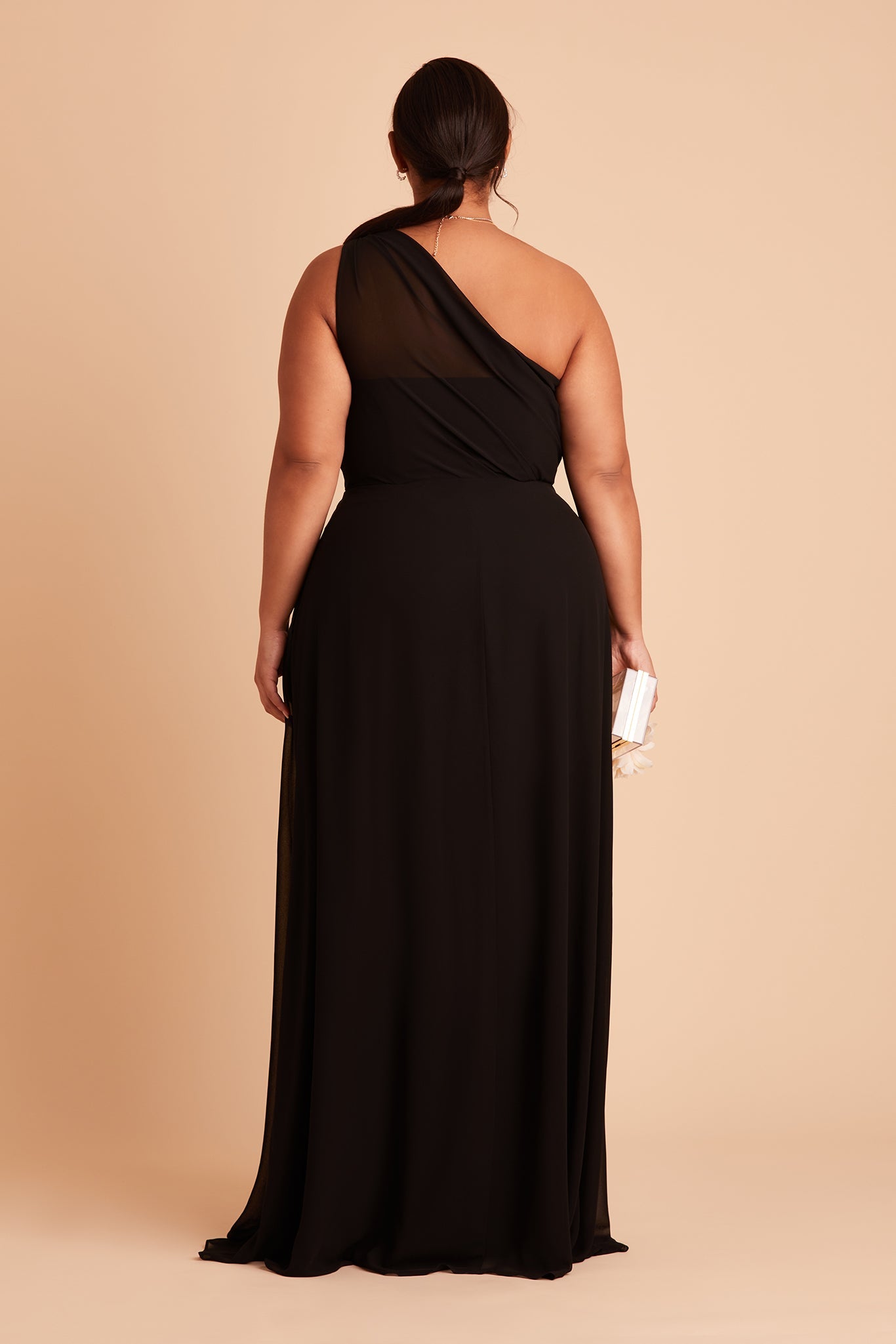 Back view of the Kira Dress Curve in black chiffon shows a full-figured model with a medium skin tone wearing sheer, softly pleating chiffon gathered at the bodice shoulder and draping across the back to the side seam as the dress flows to the floor.