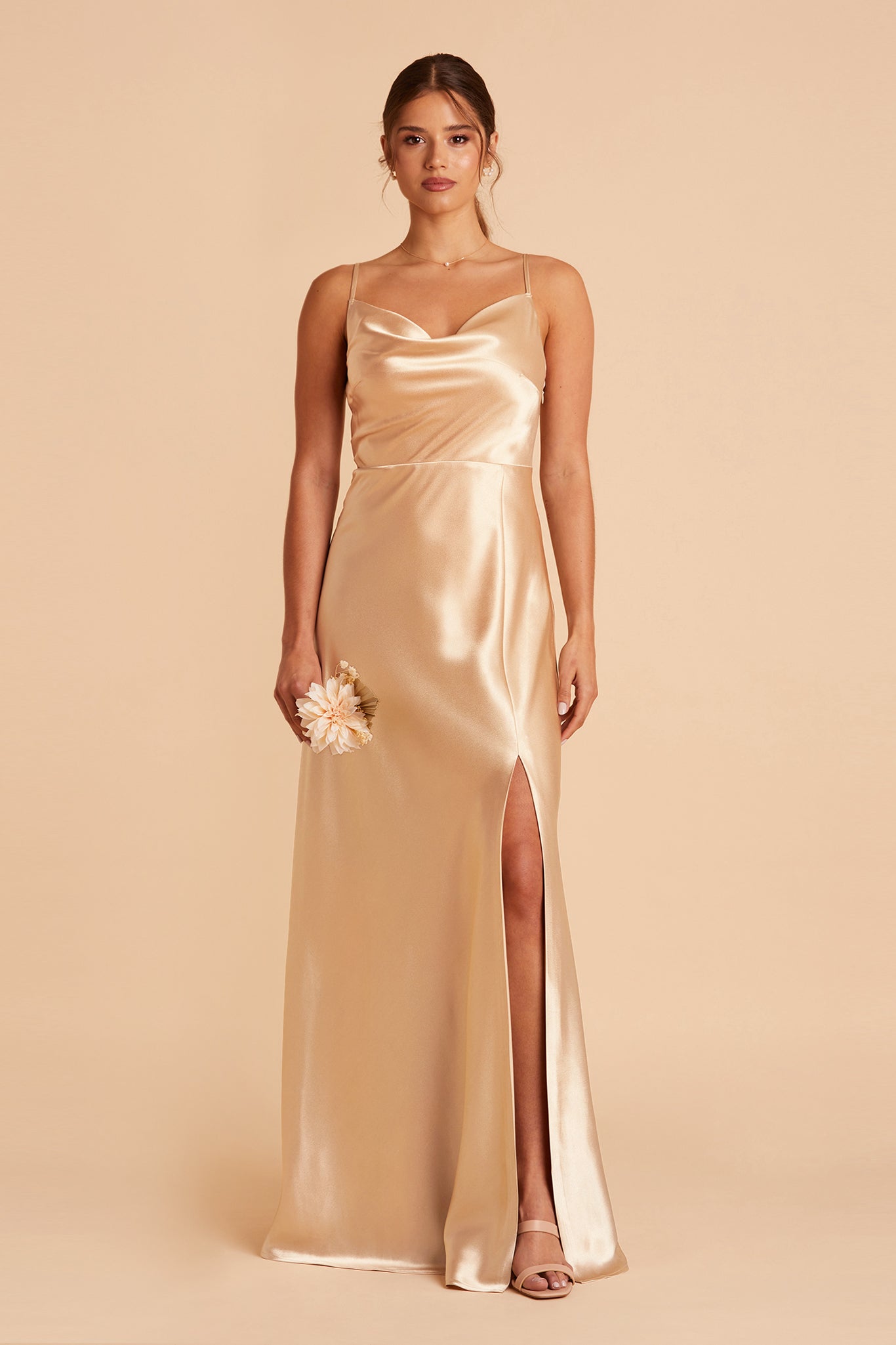 Front view of the Lisa Long Dress in gold satin shows a slender model with a light skin tone wearing a lightly draped cowl neck bodice with spaghetti straps and a floor-length flared dress with a slit. 