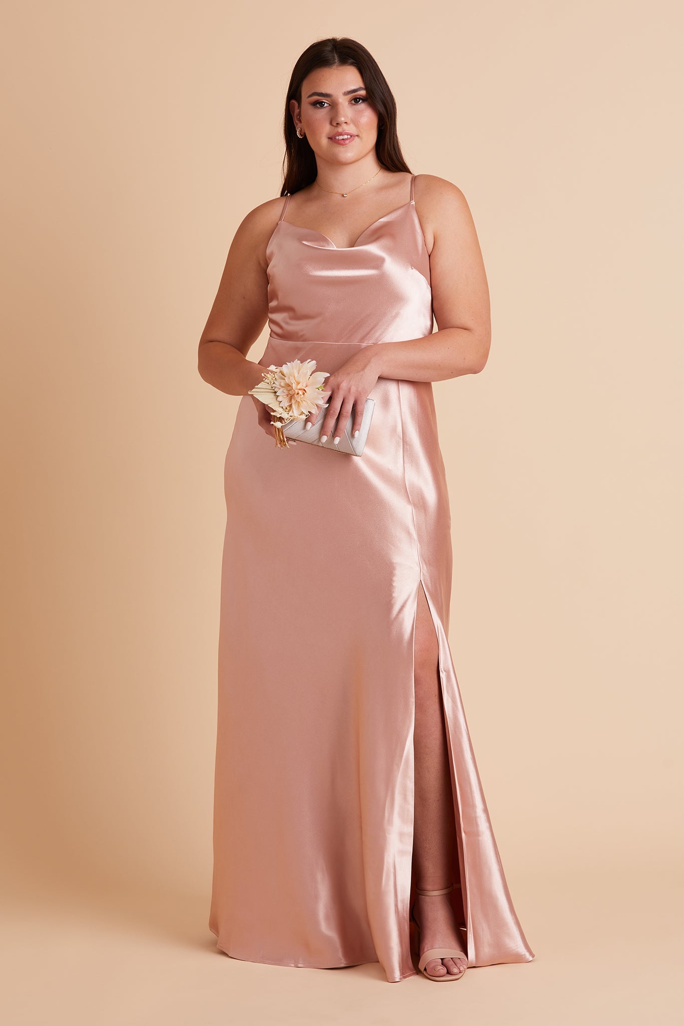 Front view of the Lisa Long Dress Curve in rose gold satin with a slit shows a full-figured model with a light skin tone wearing a lightly draped cowl neck bodice with spaghetti straps and a floor length flared dress with a slit. 