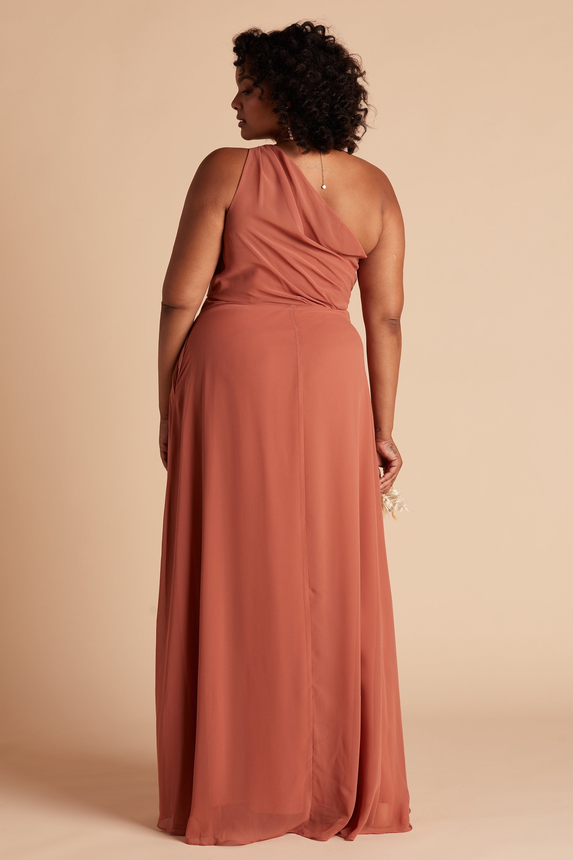Back view of the Kira Dress Curve in terracotta chiffon shows a full-figured model with a medium skin tone. The back of the dress has sheer chiffon fabric which creates a pleated Grecian one-shoulder neckline and bodice. 