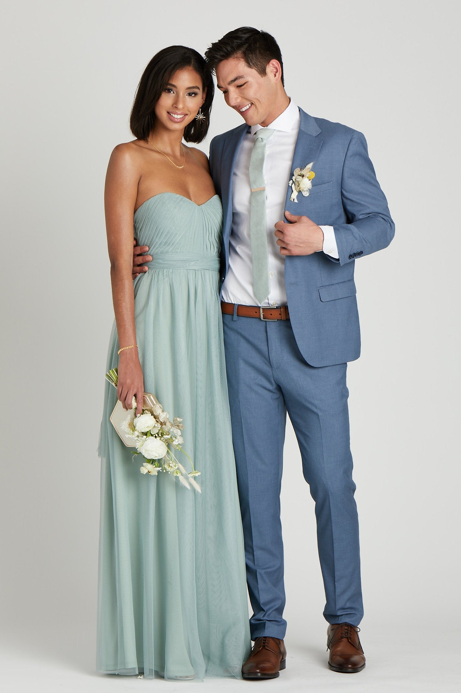 Front view  of two models in a side embrace at the waist. One model with a slender physique and a medium skin tone wears the Simon Necktie in sage with a white collared button down shirt and a medium blue suit jacket and slacks. The second model with a slender figure and a medium skin tone coordinates their look wearing a floor length strapless dress in sage. 