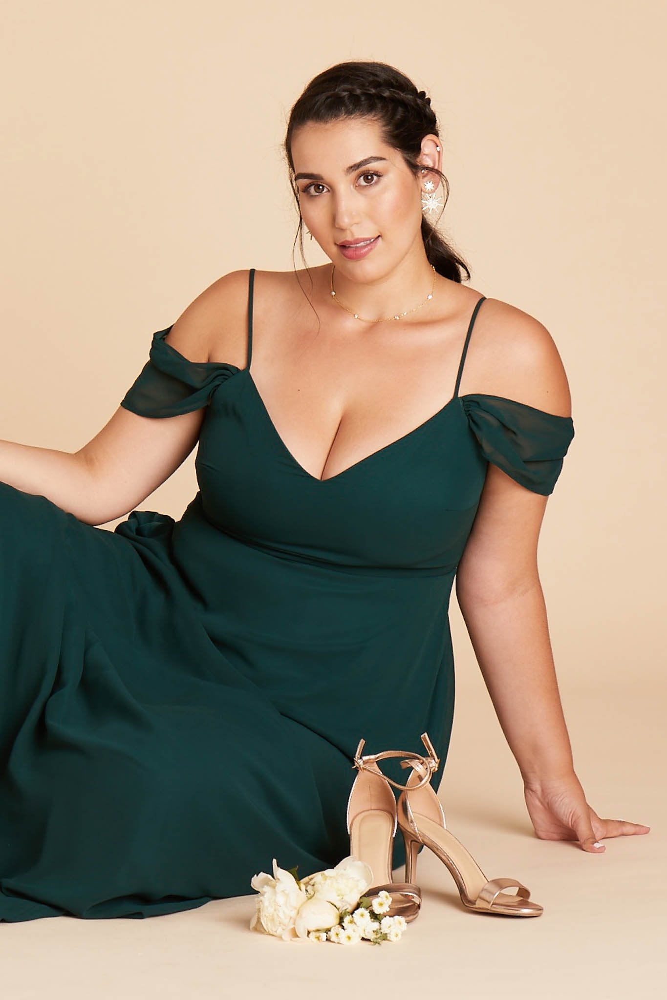 Front view of a curvy model wearing the Devin Convertible Plus Size Bridesmaid Dress in emerald chiffon sitting on the floor next to a pair of Jenny Stiletto Heels in gold. 