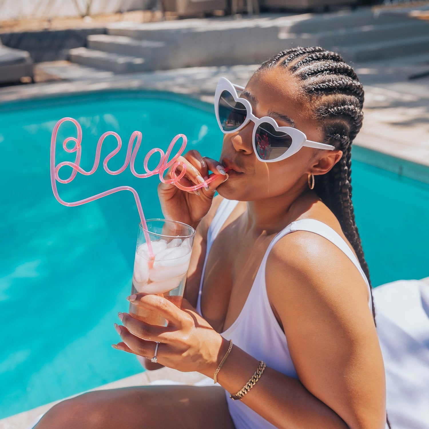 Front view of a slender model with dark skin wearing the Heart Sunglasses in white next to the pool while she sips from a silly straw that is shaped like the word “Bride.”