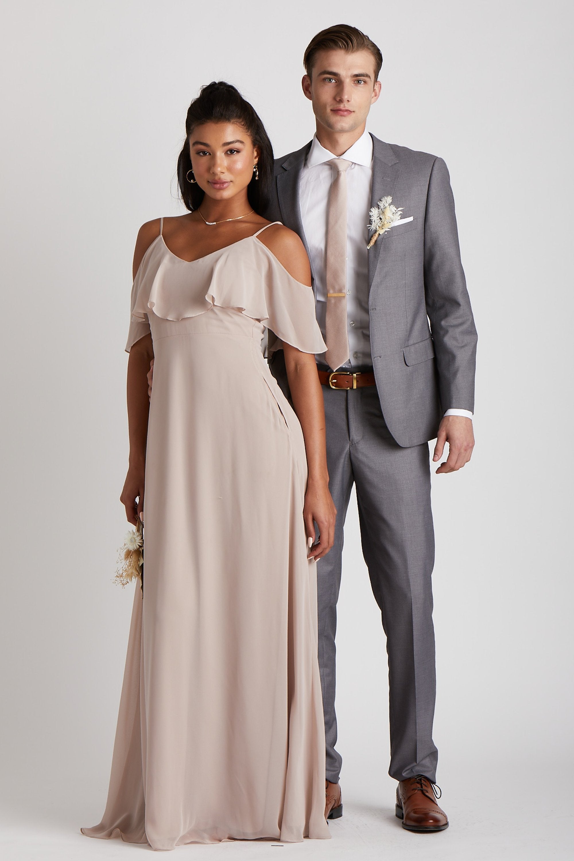 Front view  of two models standing next to each other in a staggered pose. One model has a slender physique and a light skin tone and wears the Simon Necktie in taupe with a white collared button down shirt and a medium grey suit. The other model has a slender figure and a medium skin tone and coordinates their look wearing the Jane's Convertible Dress chiffon in taupe. 