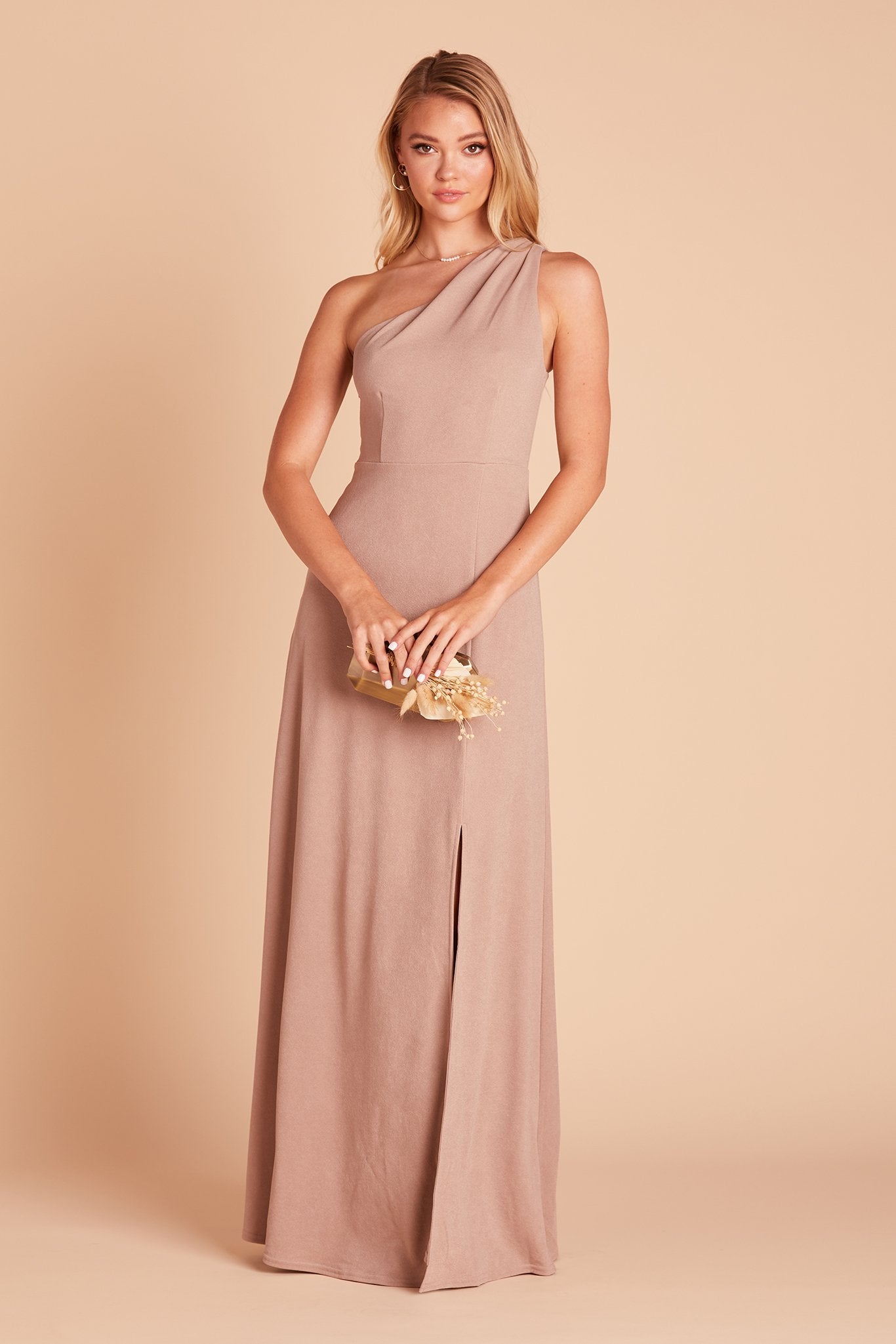 Front view of the Kira Dress in taupe crepe shows a slender model with a light skin tone wearing an asymmetrical one-shoulder, full-length dress. Soft pleating gathers at the shoulder of the bodice with a smooth fit at the waist as the dress skirt with a slight A-line silhouette flows to the floor. 
