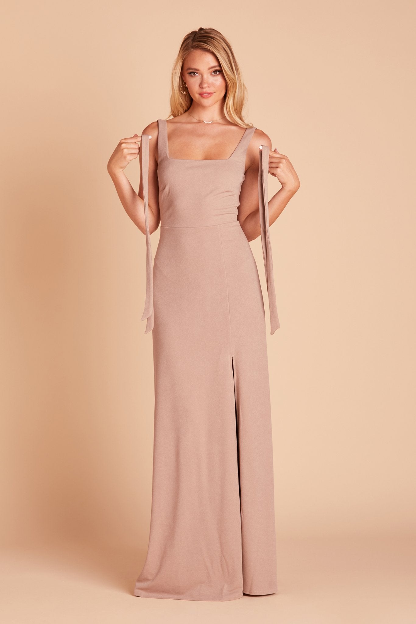 Front view of the Alex Convertible Dress in taupe as the model holds up the removable shoulder ties in each hand, letting them drape down to show their length from shoulder to hip height. 