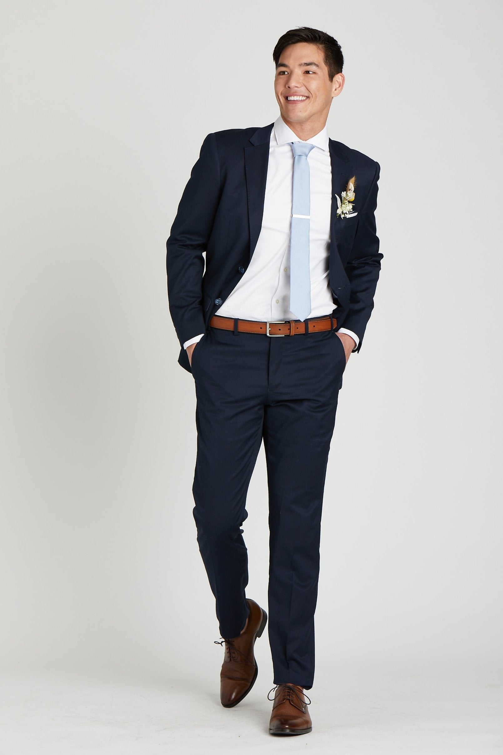 Front full view of a model wearing the Simon Necktie in dusty blue coordinated with a navy blue suit with white collared shirt, medium brown shoes and belt, white pocket square, and a dried flower boutonniere in ivory.