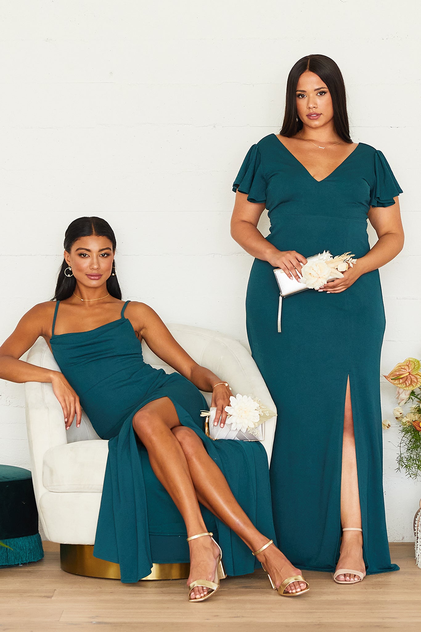 Two models pose in Birdy Grey emerald bridesmaid dresses. On the left, a slender model with a medium skin tone lounges in a white chair wearing the Ash Bridesmaid Dress. On the right, a curvy model with a medium skin tone stands next to the chair, wearing the floor-length Hannah Empire Curve Dress with Flutter Sleeves.