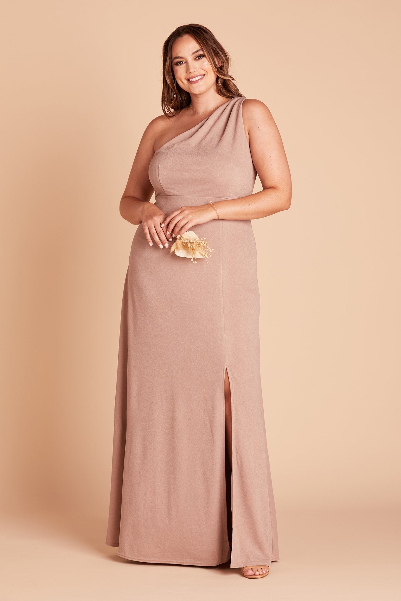 Front view of the Kira Dress Curve in taupe crepe with optional slit shows a full-figured model with a light skin tone wearing an asymmetrical one-shoulder, full-length dress. Soft pleating gathers at the left shoulder of the bodice with a smooth fit at the waist as the dress skirt with a slight A-line silhouette flows to the floor.   
