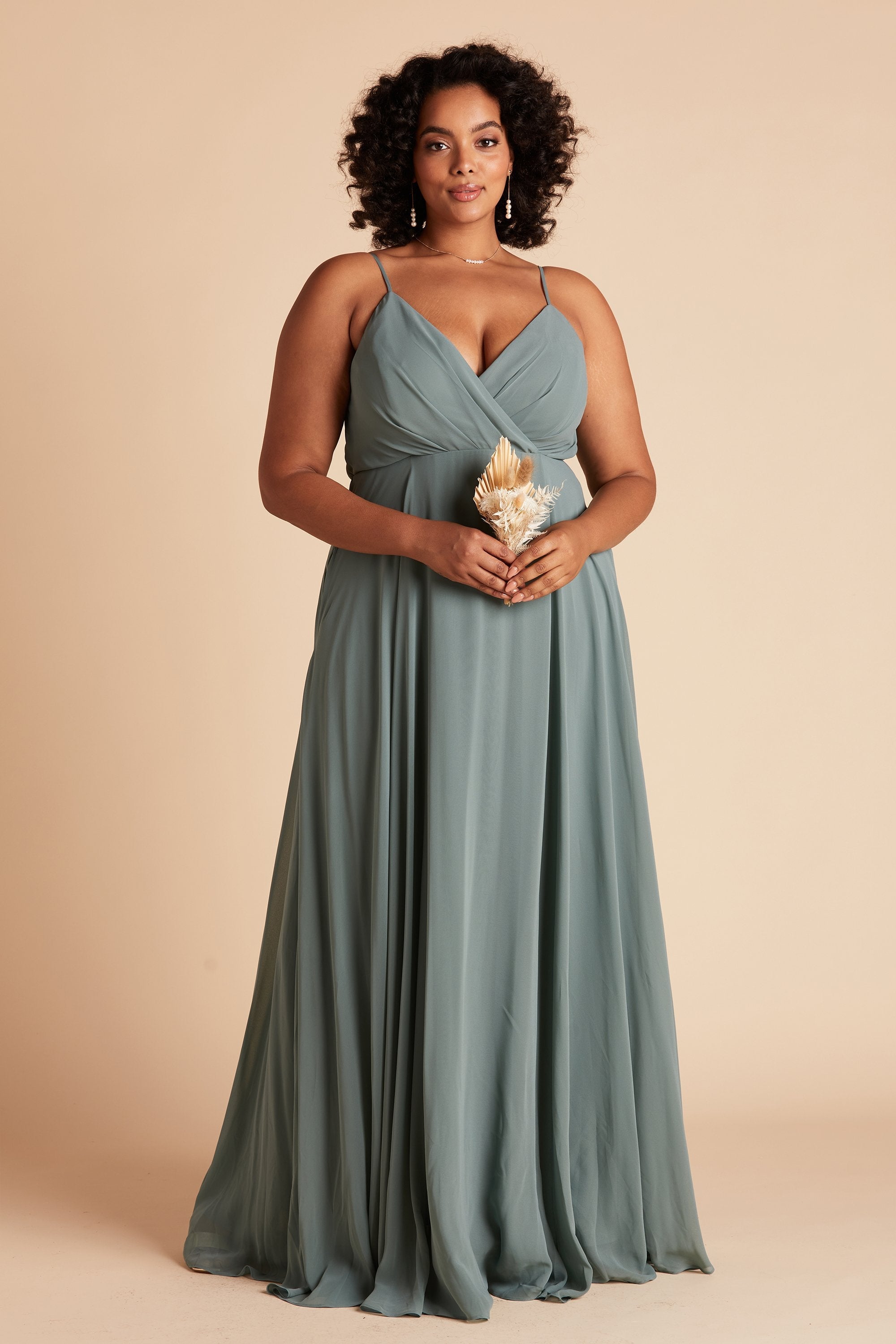 Front view of the floor-length Kaia Dress Curve in sea glass chiffon shows a full-figured model with a medium skin tone wearing a spaghetti strap V-neck dress with a raised empire waistline wraps across their full bosom showing cleavage.