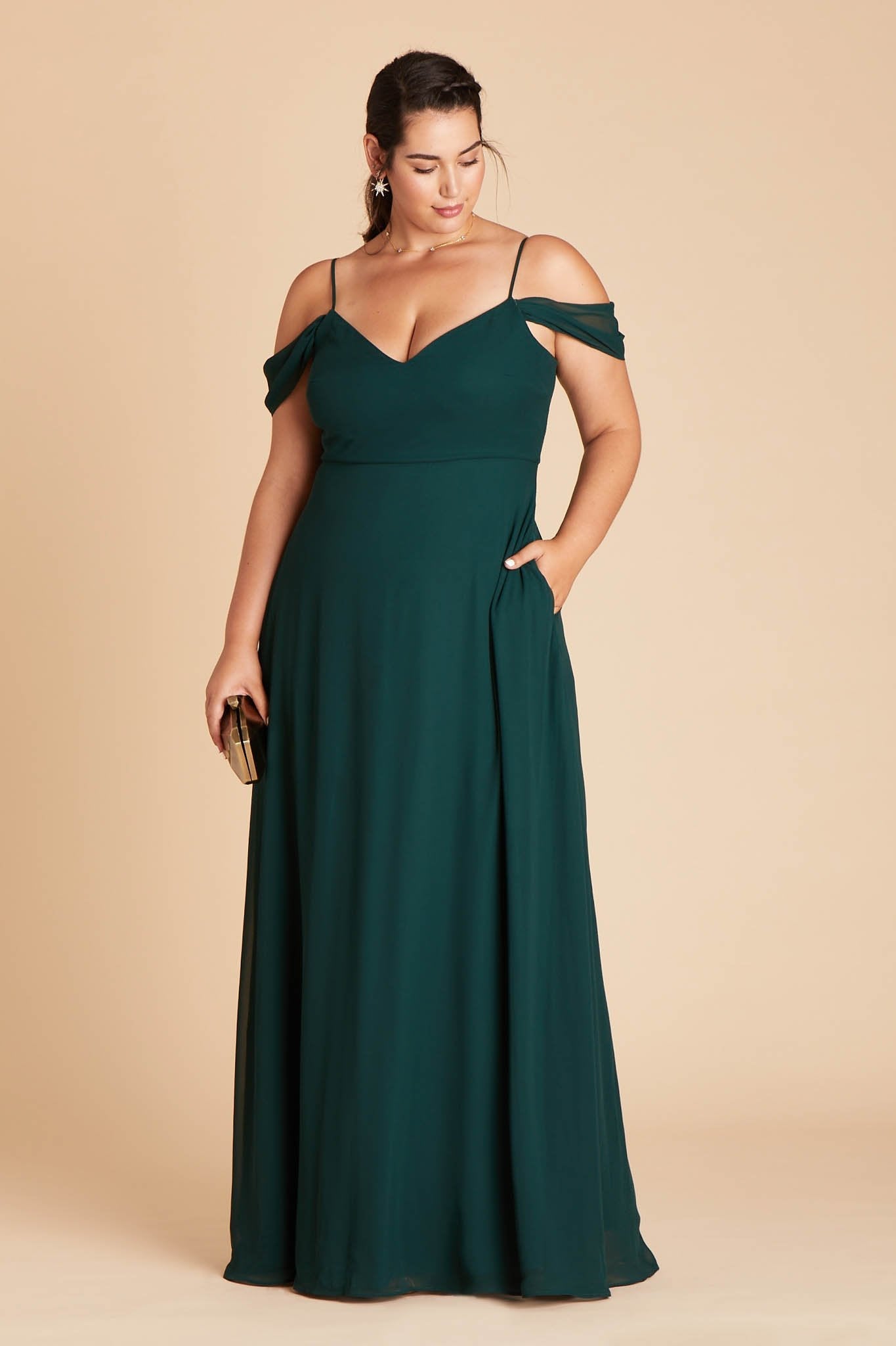Front view of the floor-length Devin Convertible Plus Size Bridesmaid Dress in emerald chiffon worn by a curvy model with a light skin tone. The model rests her left hand in the hidden side pocket.
