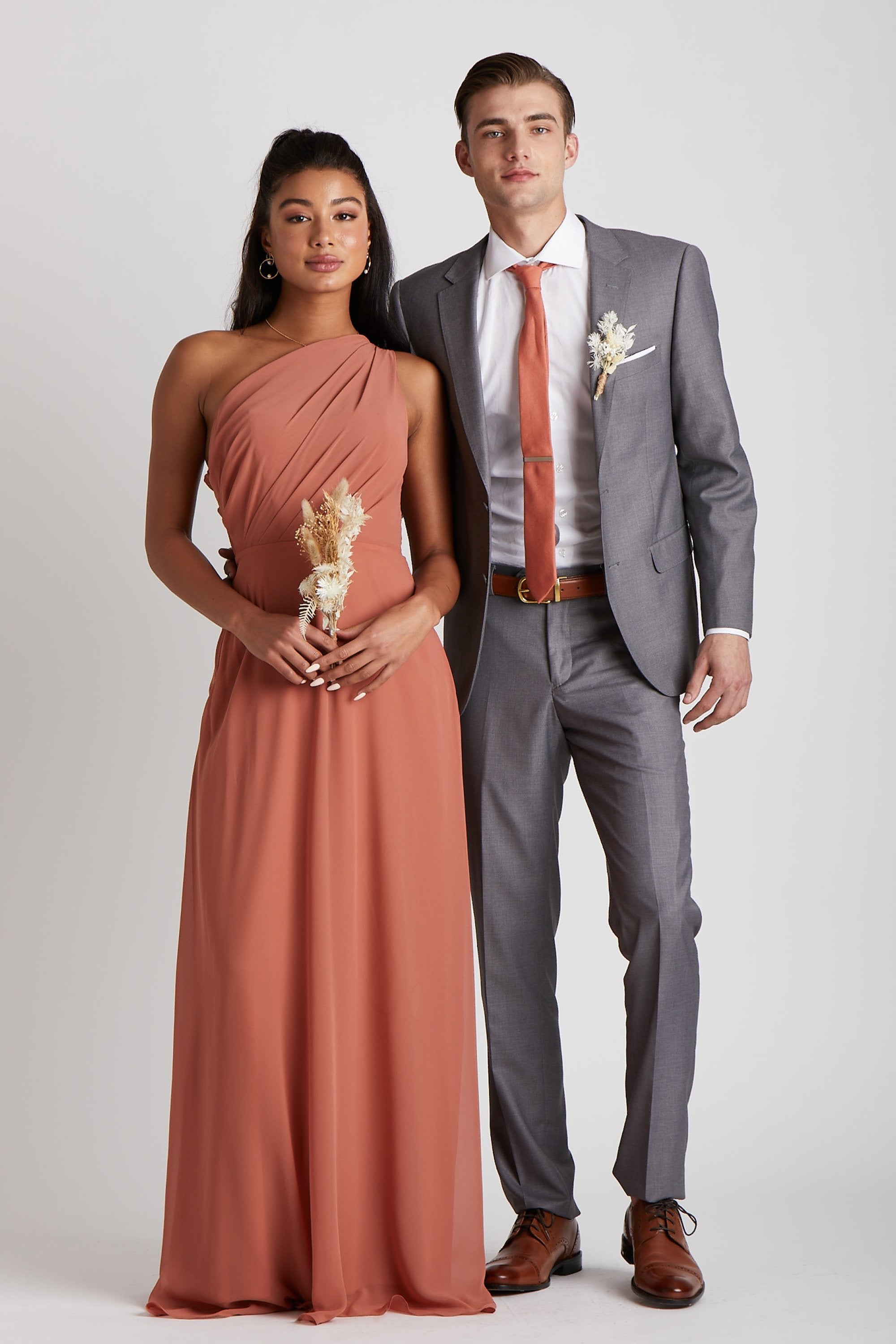 Front view of two models standing next to each other. The model on the right has a slender physique and a light skin tone and wears the Simon Necktie in terracotta with a white collared button down shirt and a medium grey suit. The model in front has a slender figure and a medium skin tone coordinating their look wearing the Kira Dress in terracotta chiffon.