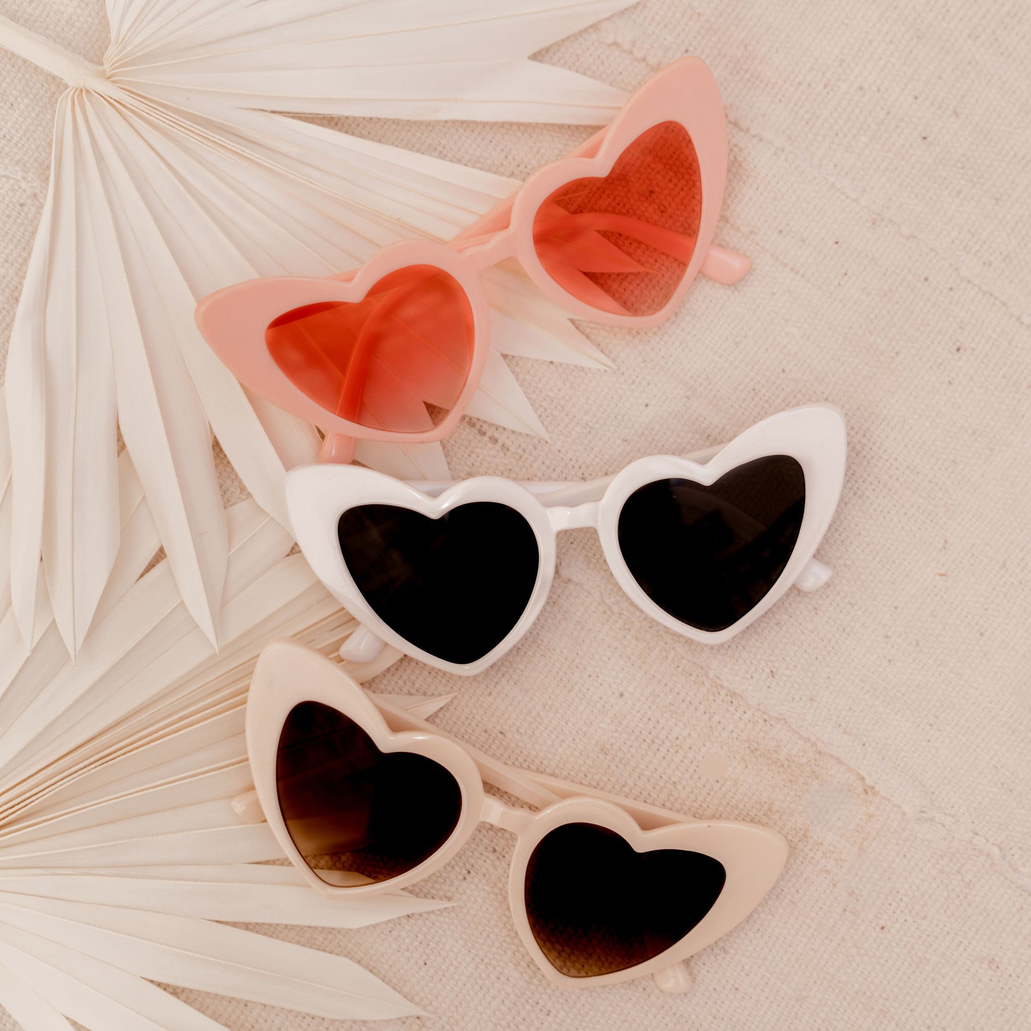 From above, we see three pairs of Heart Sunglasses by Birdy Grey next to two handheld fans. The first pair is pink with pink lenses. The second pair is white with black lenses. The third pair is nude with smoky lenses.