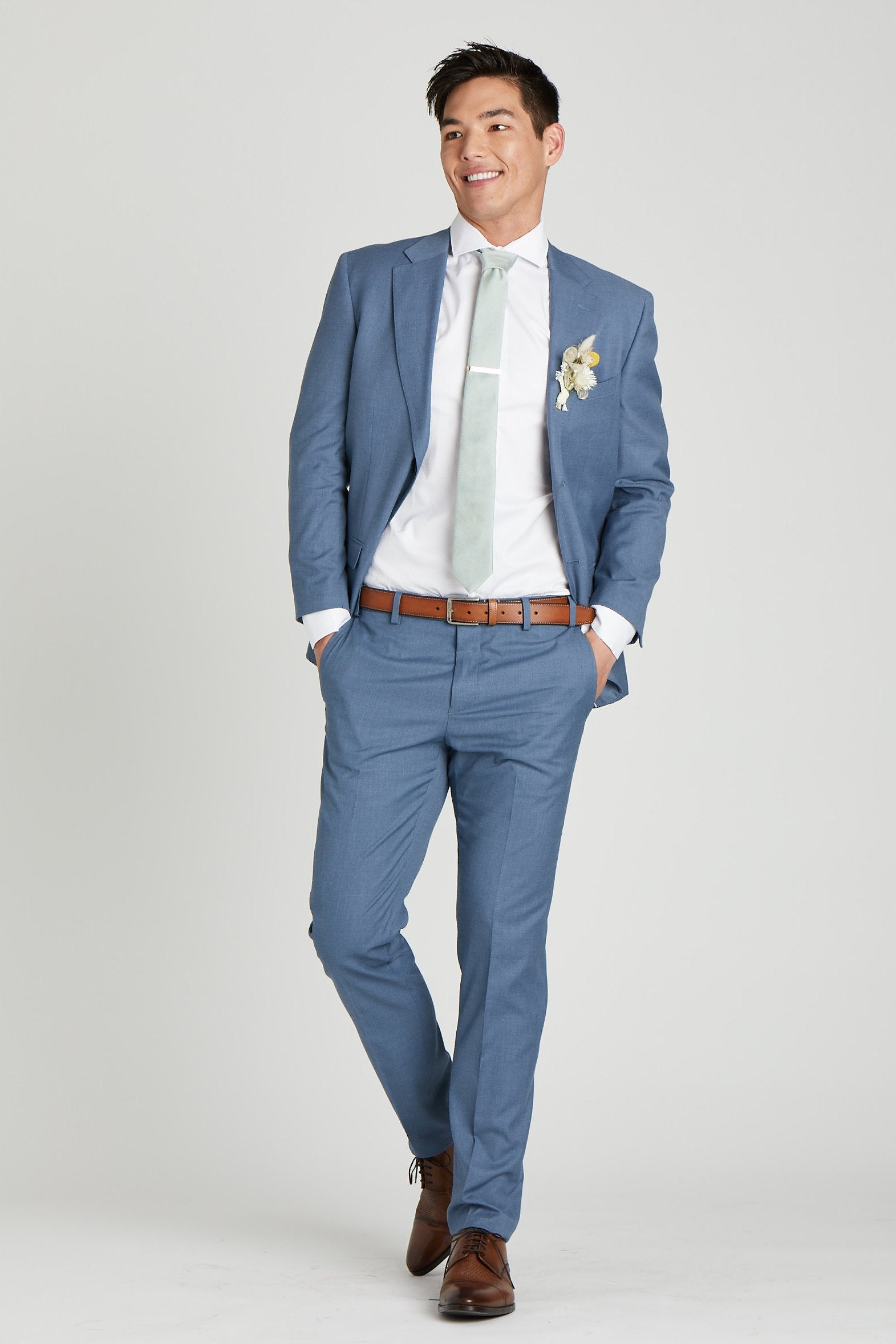 Front view of a model wearing the Simon Necktie in sage coordinated with a medium blue suit with white collared shirt, medium brown shoes and belt, and a dried flower boutonniere in white. 