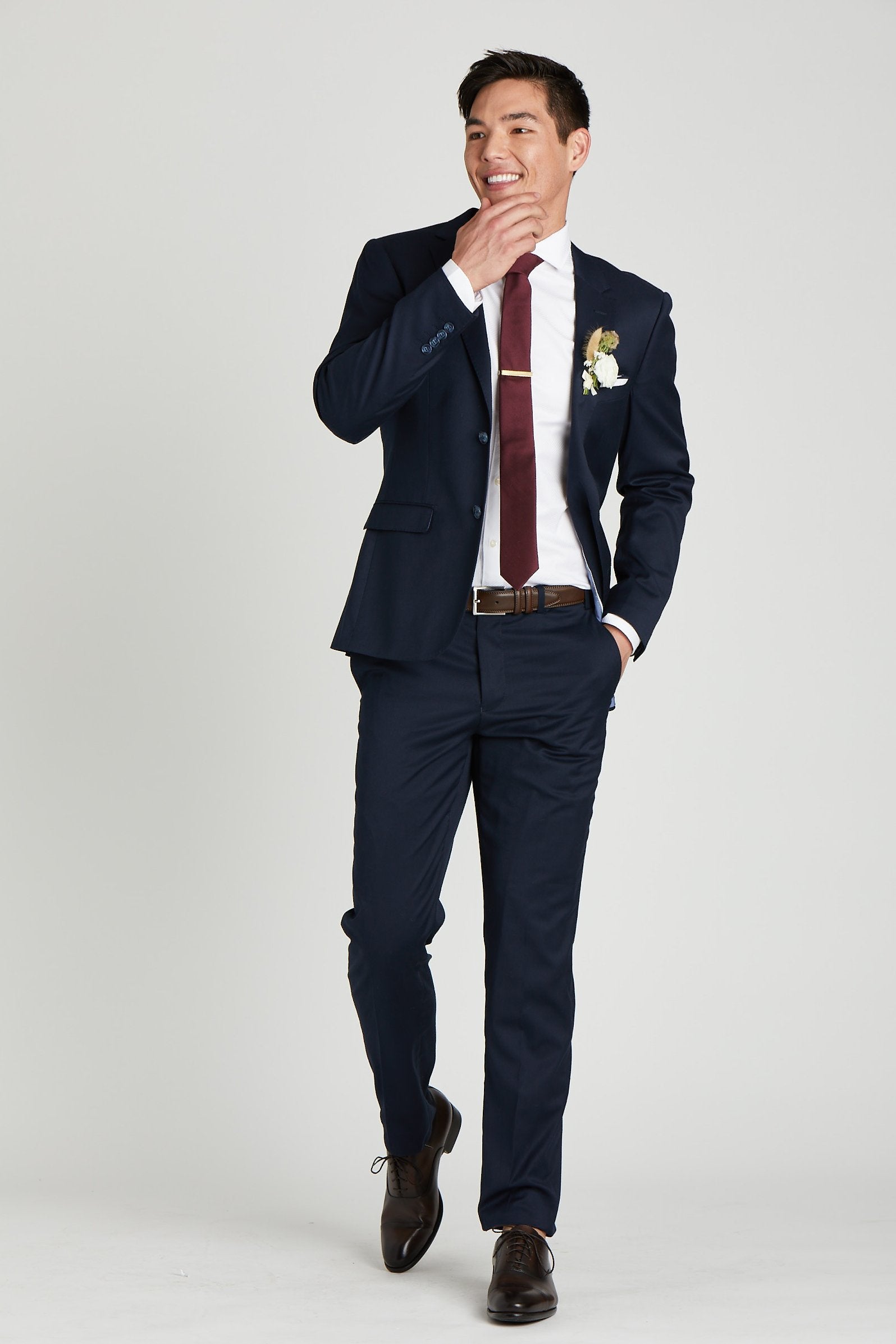 Front view of a model wearing the Simon Necktie in cabernet coordinated with a navy blue suit with white collared shirt, dark brown shoes and belt, white pocket square, and a dried flower boutonniere in white. 