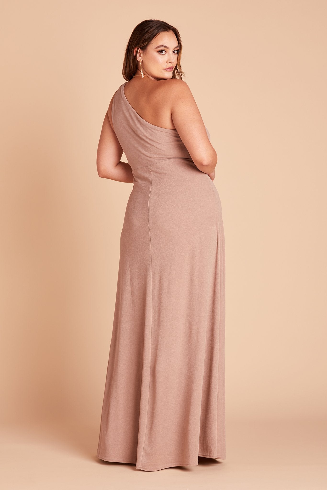 Back side view of the floor-length Kira Dress Curve in taupe crepe with a model showing soft pleating gathered at the back bodice shoulder and a smooth, conformed fit at the waist.