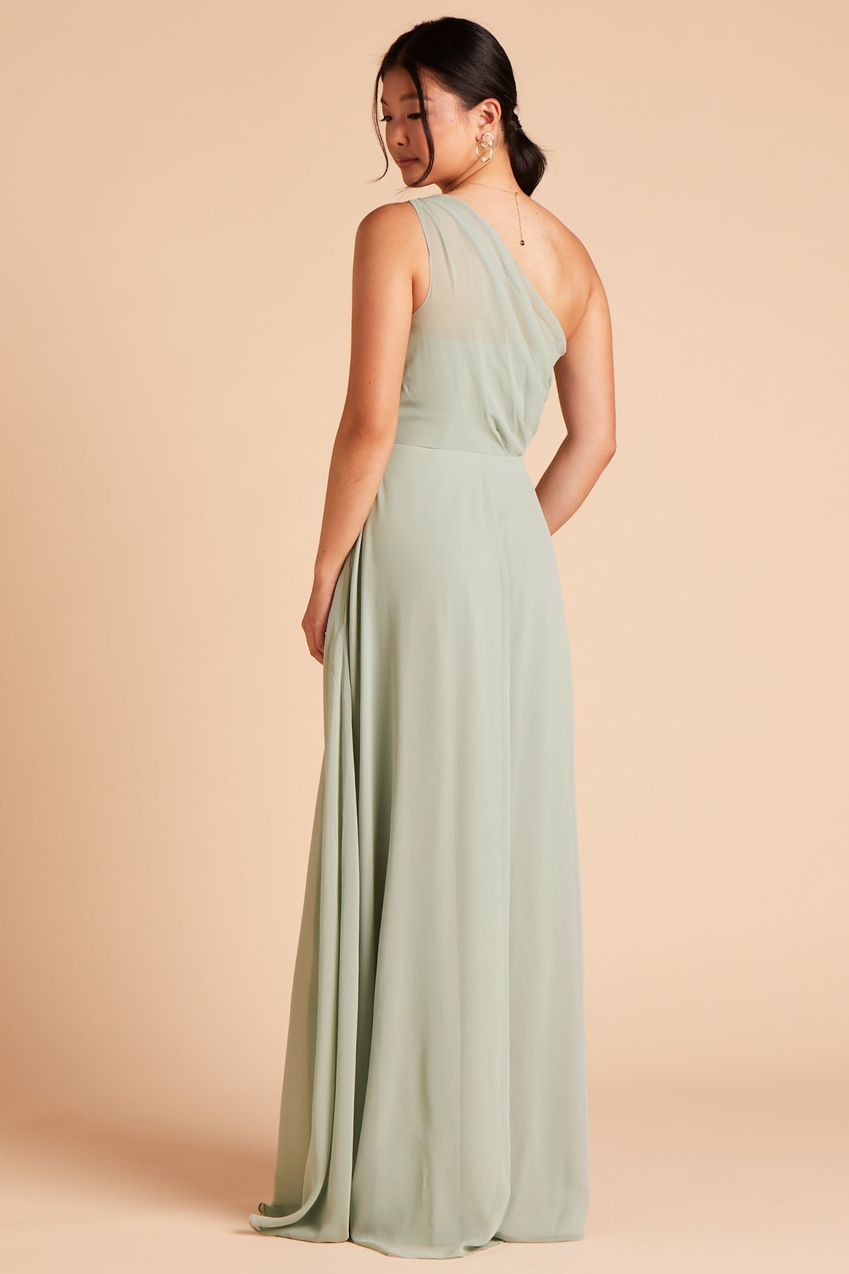 Back view of the floor-length Kira Dress in sage chiffon shows a model wearing sheer, softly pleating chiffon gathered at the bodice shoulder and draping across the back to the side seam.