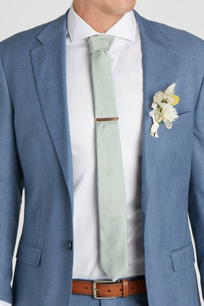 Front closeup view of a model wearing the Simon Necktie in sage with a white button down collared shirt and medium blue suit. The tie is kept in place with a narrow rectangular necktie clasp in gold.
