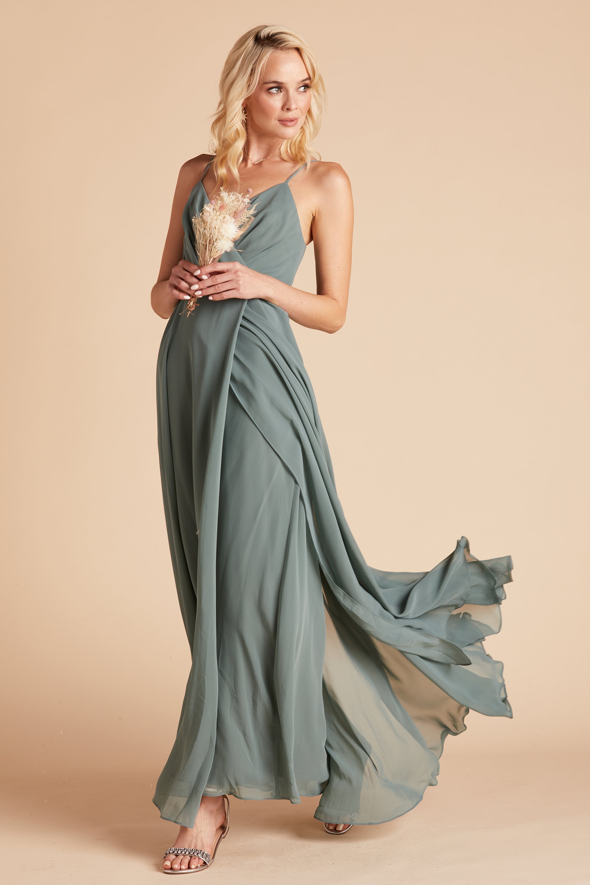 Front view of the Kaia Dress in sea glass chiffon shows a slender model with a light skin tone wearing a spaghetti strap V-neck dress that wraps across the chest to the raised empire waistline. The floor length overlapping skirt flows back behind the walking model.