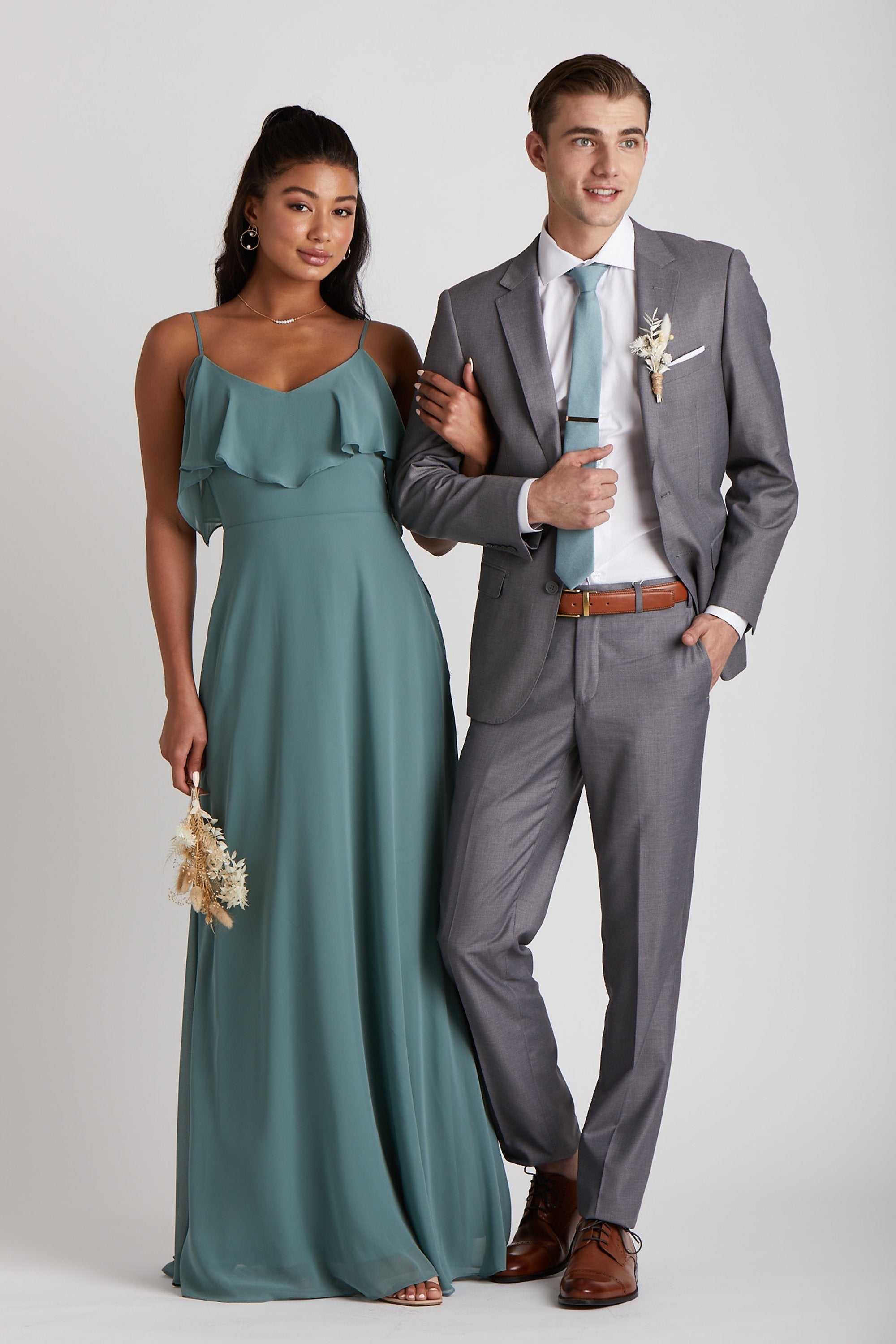 Front view of two models next to each other. One model with a slender physique and a light skin tone wears the Simon Necktie in sea glass, a white collared button down shirt, and a medium grey suit jacket and slacks. The second model with a slender figure and a medium skin tone coordinates their look wearing the Jane's Convertible Dress chiffon in sea glass. 
