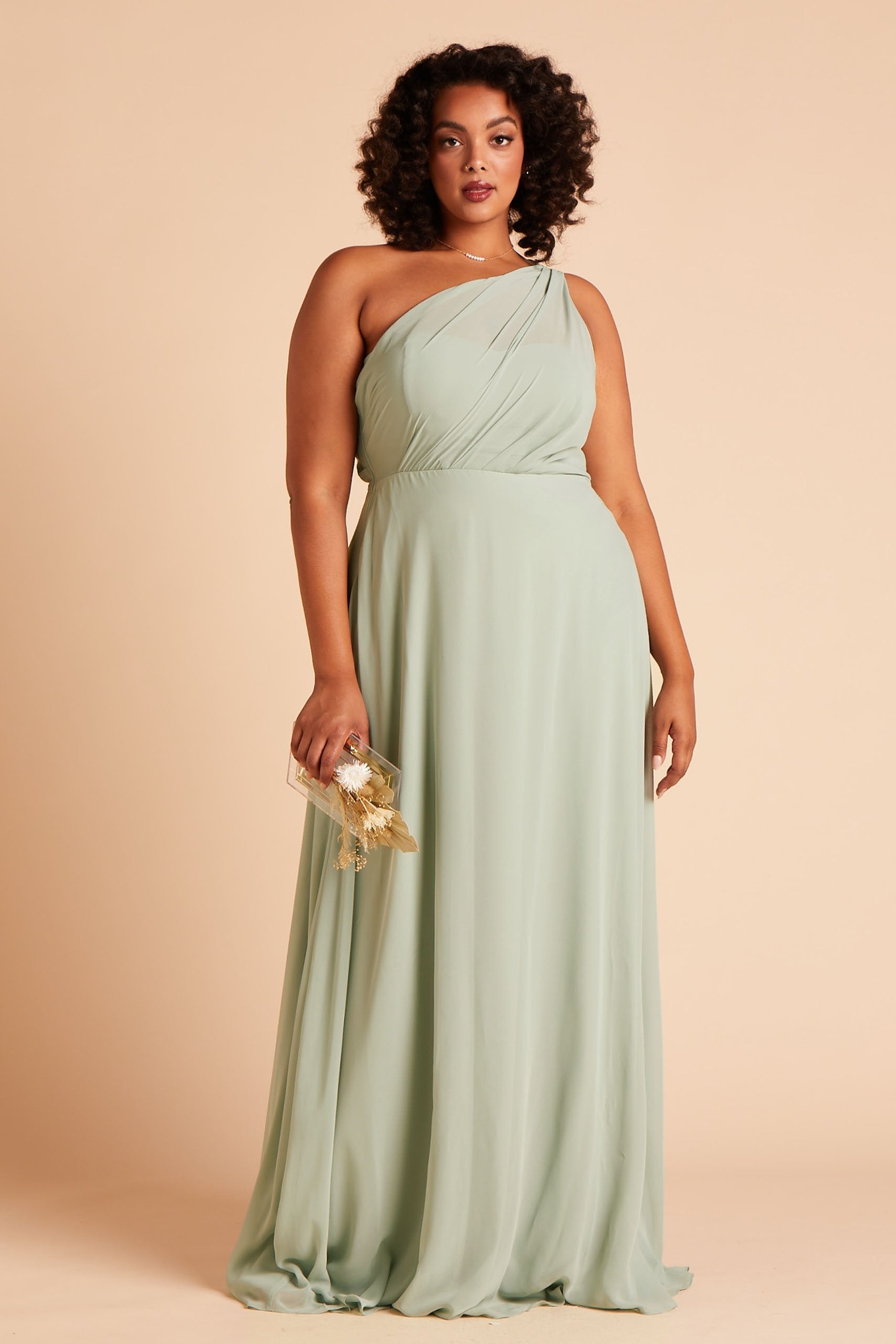 Front view of the Kira Dress Curve in sage chiffon without the optional slit shows a full-figured model with a medium skin tone holding the Clear Clutch with Clear Beads purse at their side.