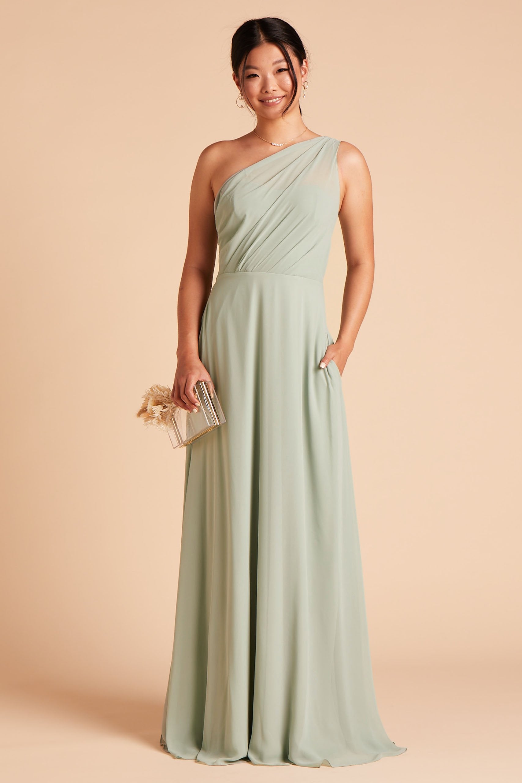 Front view of the Kira Dress in sage chiffon without the optional slit shows a slender model with a medium skin tone wearing an asymmetrical one-shoulder, full-length dress. Soft pleating gathers at the left shoulder of the bodice with a smooth fit at the waist as the dress skirt with a slight A-line silhouette flows to the floor.