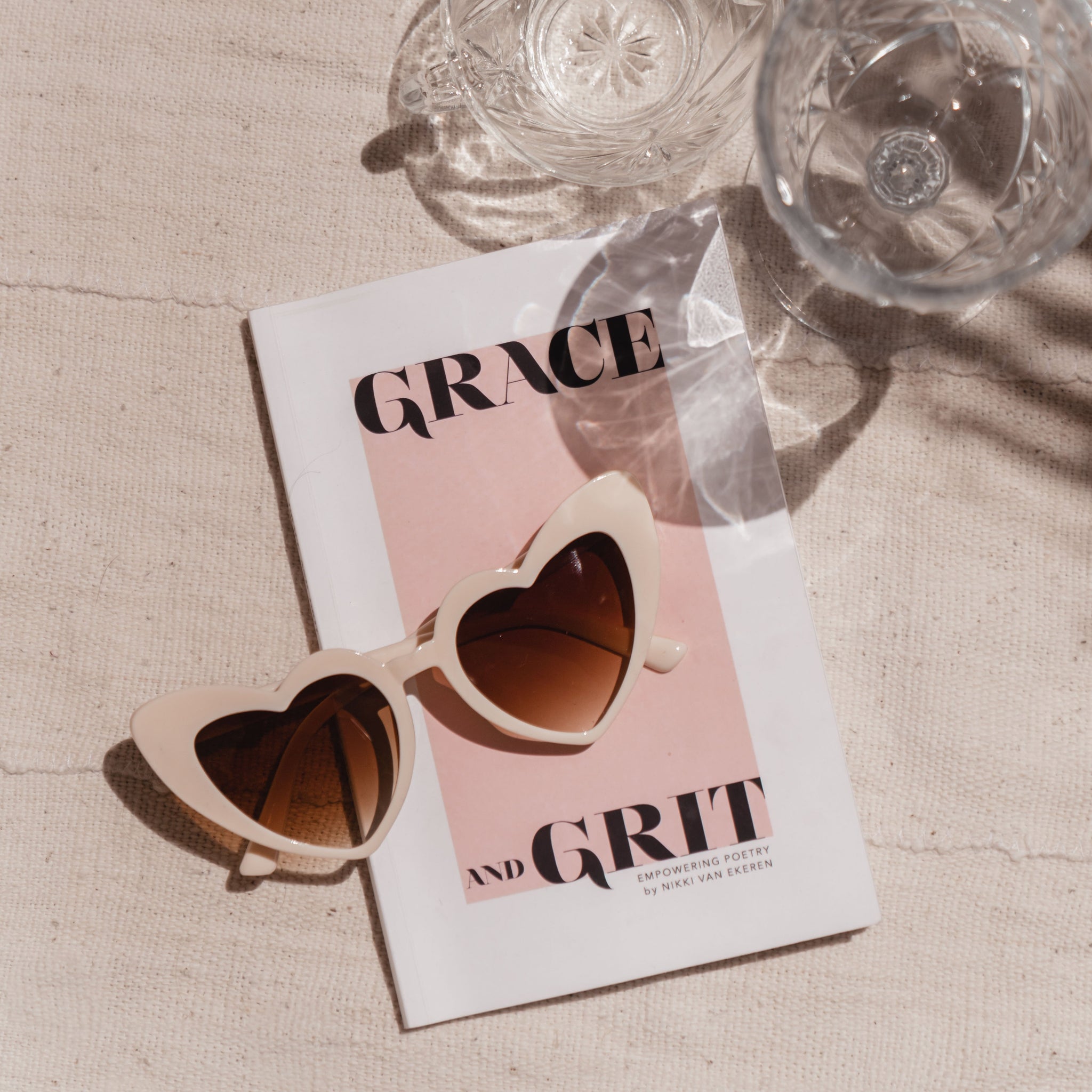 From above, the Heart Sunglasses by Birdy Grey in nude lay atop a pink and white book titled, “Grace and Grit: Empowering Poetry by Nikki Van Ekeren.”
