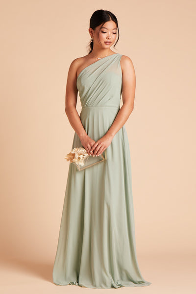 Front view of the Kira Dress in sage chiffon without the optional slit shows a model wearing the Brookhaven Pearl Trio Necklace as they hold the Clear Clutch with Clear Beads purse at their side.
