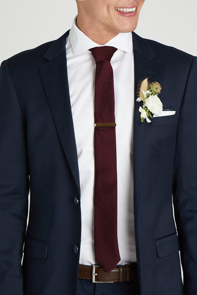 Front closeup view of the model wearing the Simon Necktie in cabernet with a white button down collared shirt and navy blue suit. The tie is kept in place with a narrow rectangular tie clasp in matte gold.