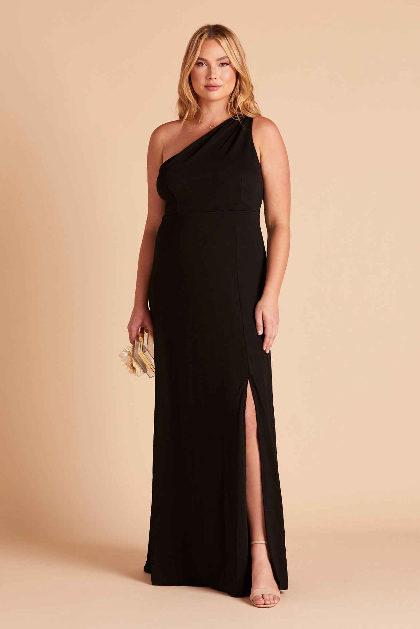 Front view of the Kira Dress in black crepe has a curvy model showing the left slit above their knee. They are wearing the Elle Chunky Heel shoe in blush and hold the Clear Clutch with Clear Beads purse at their side. 