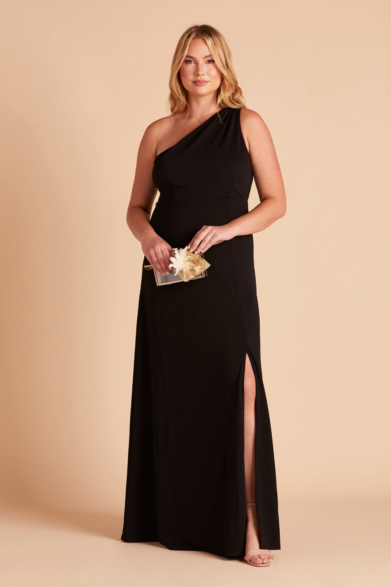 Front view of the Kira Dress Curve in black crepe with optional slit shows a curvy model with a light skin tone wearing an asymmetrical one-shoulder, floor length dress. Soft pleating gathers at the shoulder of the bodice with a smooth fit at the waist. The dress skirt with a slight A-line silhouette flows to the floor. 