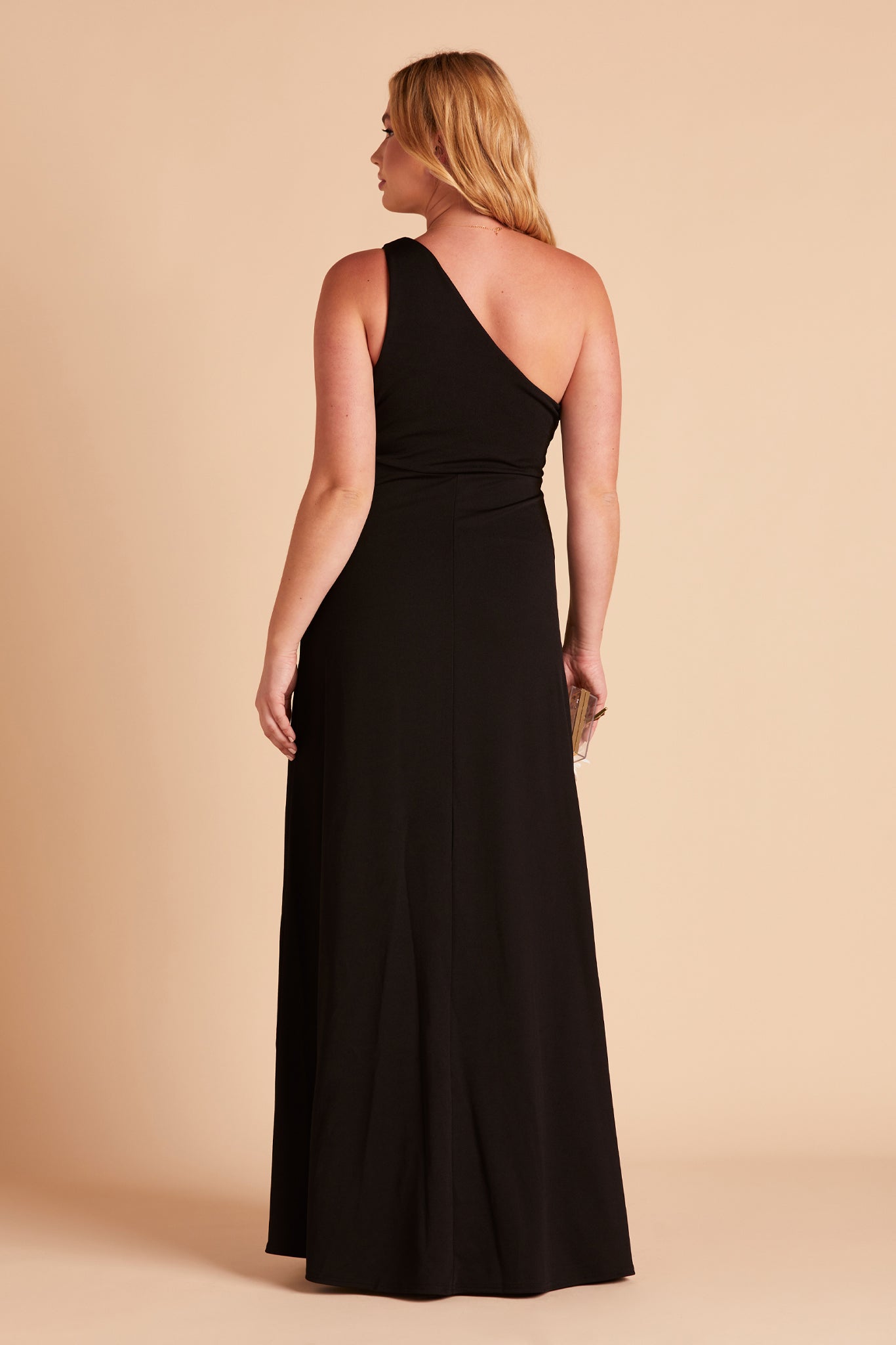 Back view of the full-length Kira Dress in black crepe shows a curvy model wearing an asymmetrical one-shoulder, full-length dress. Soft pleating gathers at the shoulder of the bodice with a smooth, conformed fit at the waist. 