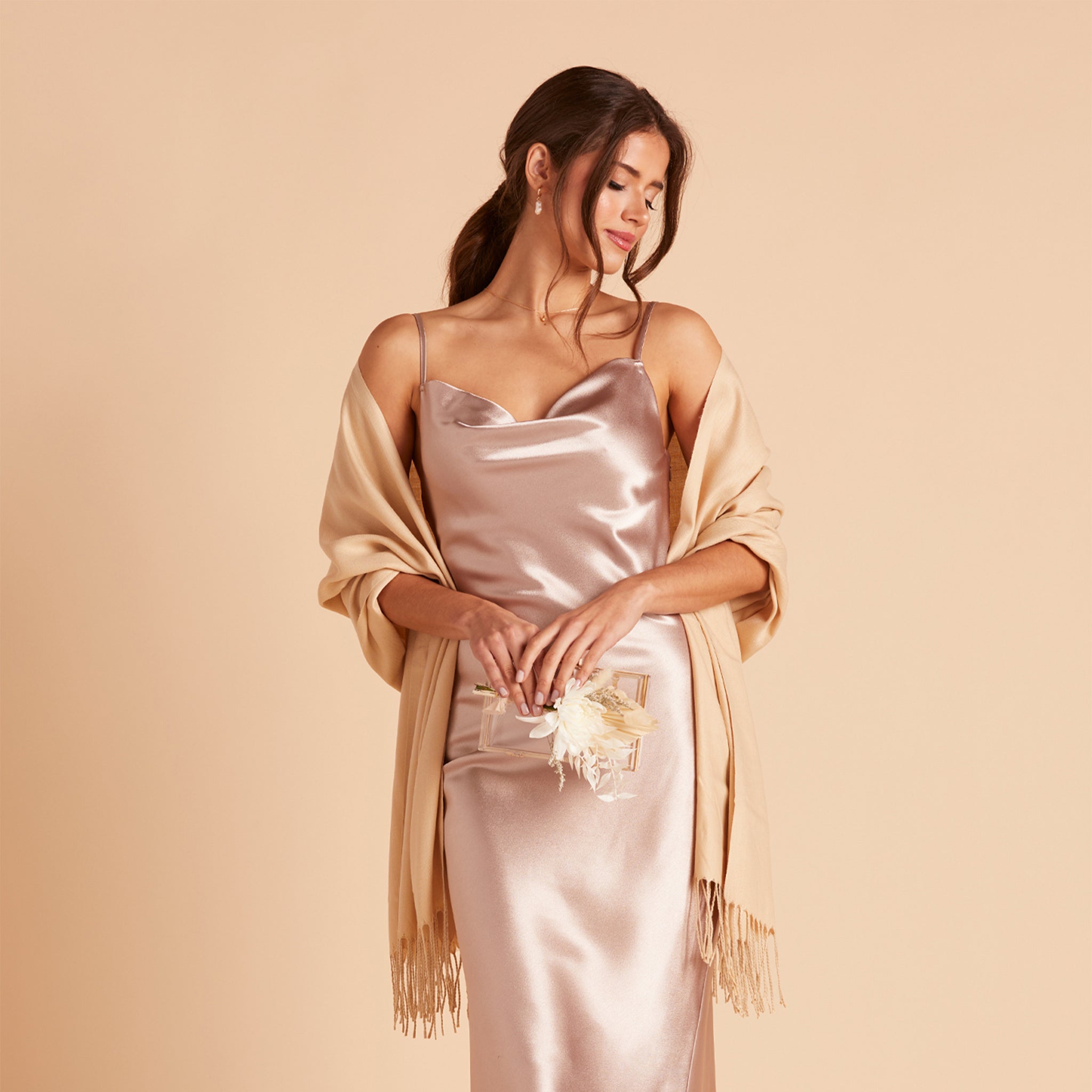 Front view of the Shawl with Fringe in beige shows a model with a slender figure and a medium skin tone wearing the Lisa Satin Midi Dress in taupe with the shawl draped across their shoulders and back and the fringe ends over their bent arms. 