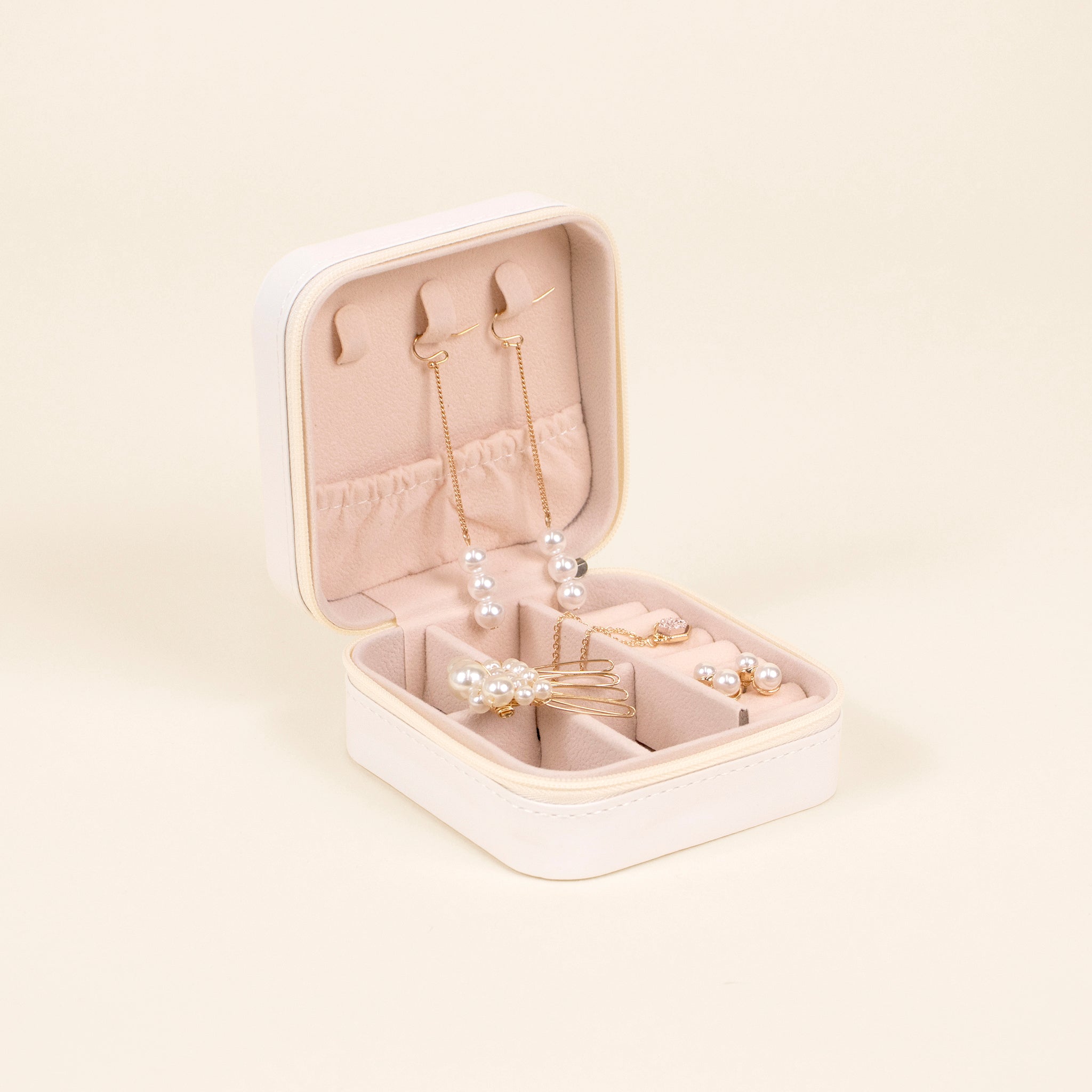 Interior view of the White Jewelry Box contains dangling and stud earrings, hair clips, and necklaces hanging from tabs or tucked into individual compartments and slots. The interior material is blush pink in color. 