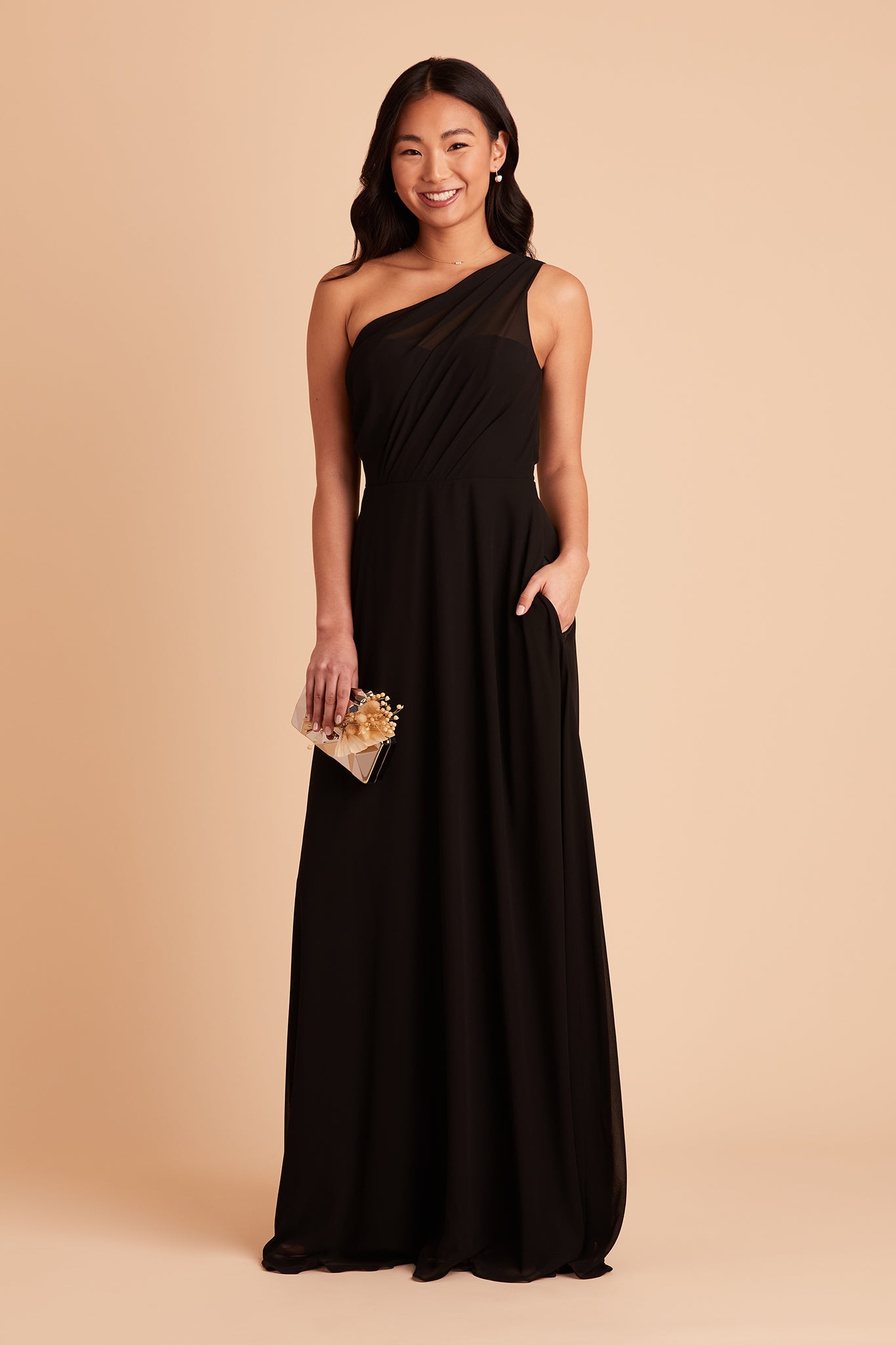 Front view of the Kira Dress in black chiffon shows a model with their hand tucked into their left pocket. 