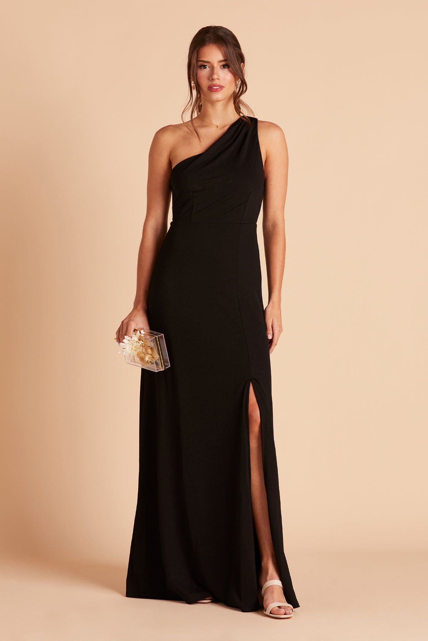 Front view of the Kira Dress in black crepe shows a slender model with a light skin tone wearing an asymmetrical one-shoulder, full-length dress. Soft pleating gathers at the shoulder of the bodice with a smooth fit at the waist as the dress skirt with a slight A-line silhouette flows to the floor. 