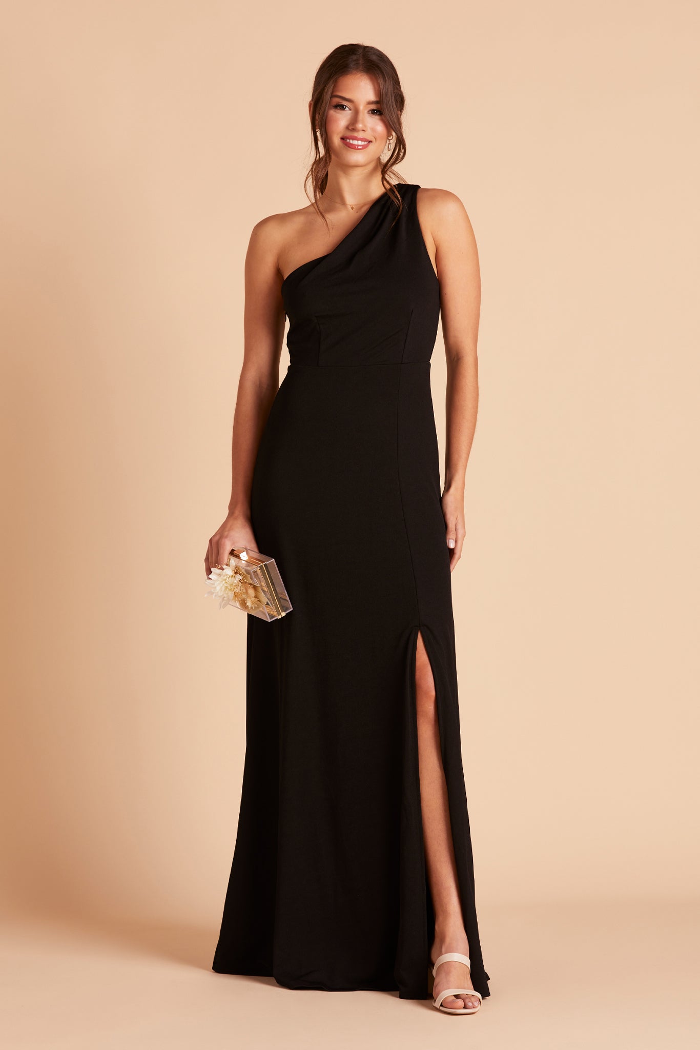 Front view of the Kira Dress in black crepe shows a model wearing a full-length dress with mid-thigh slit and leg revealed. They wear the Mary High Chunky Heel shoe in almond. 