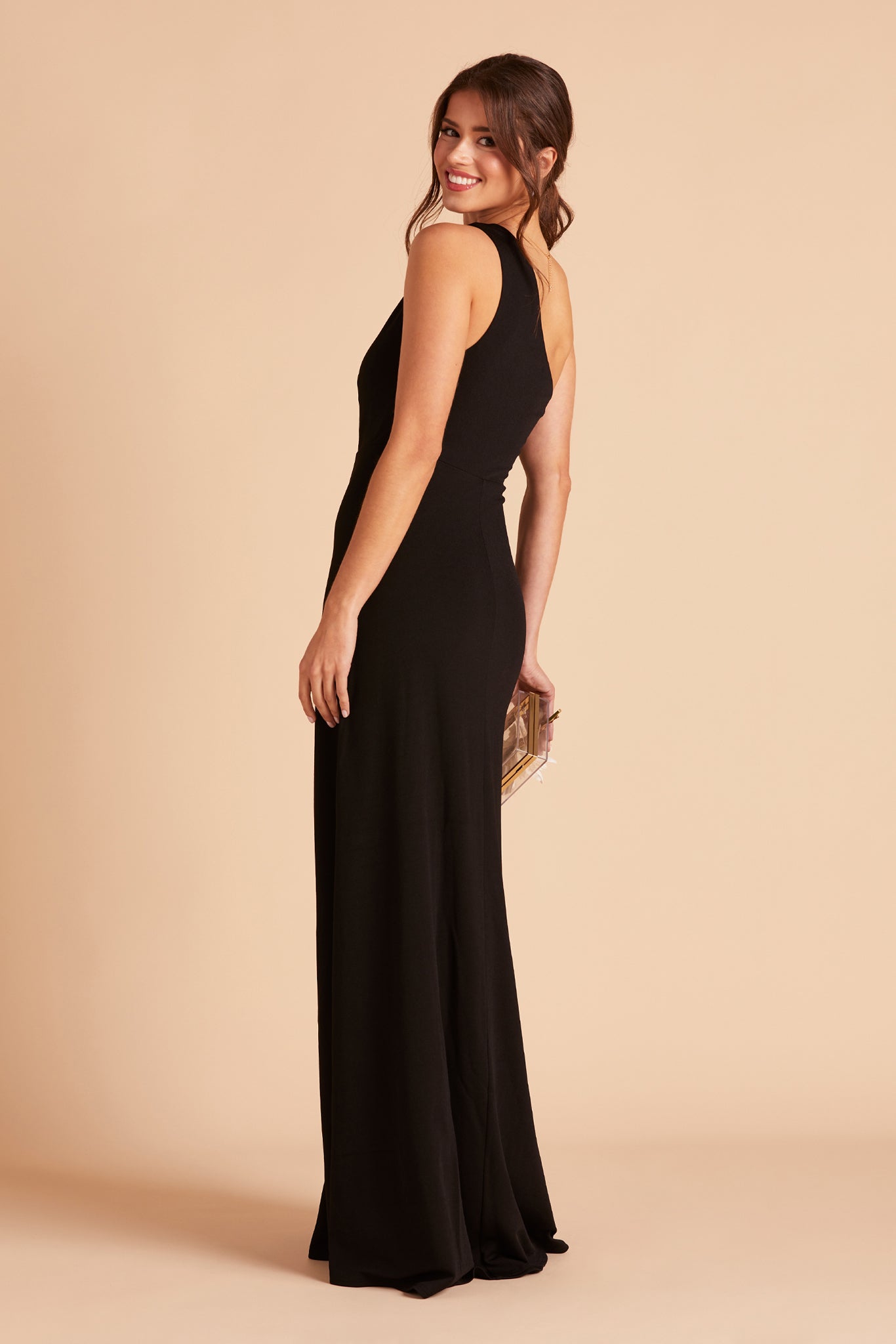 Back side view of the floor-length Kira Dress in black crepe shows a slender model with a light skin tone showing soft pleating gathered at the bodice shoulder and a smooth, conformed fit at the waist.