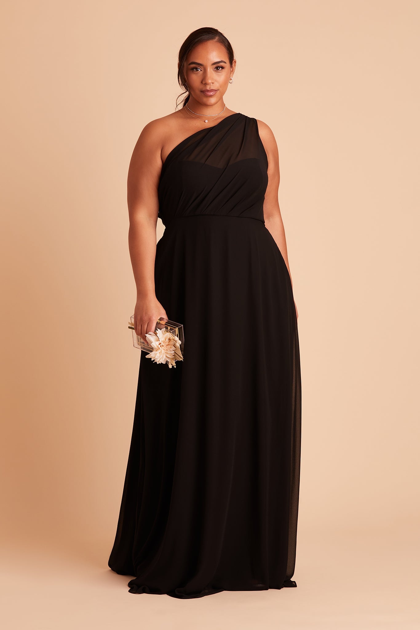 Front view of the floor-length Kira Dress Curve in black chiffon without the optional slit shows a full-figured model with a medium skin tone wearing an asymmetrical one-shoulder, full-length dress. Soft pleating gathers at the left shoulder of the bodice and drapes over their full bosom finishing with a smooth fit at the waist.