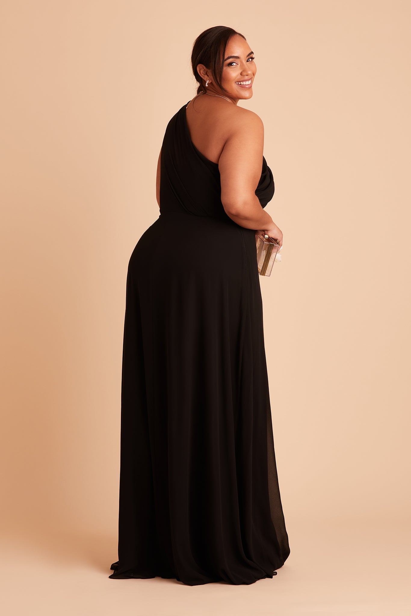 Front view of the Kira Dress Curve in black chiffon without the optional slit shows a slender model with a light skin tone wearing an asymmetrical one-shoulder, full-length dress. Soft pleating gathers at the left shoulder of the bodice with a smooth fit at the waist as the dress flows to the floor.