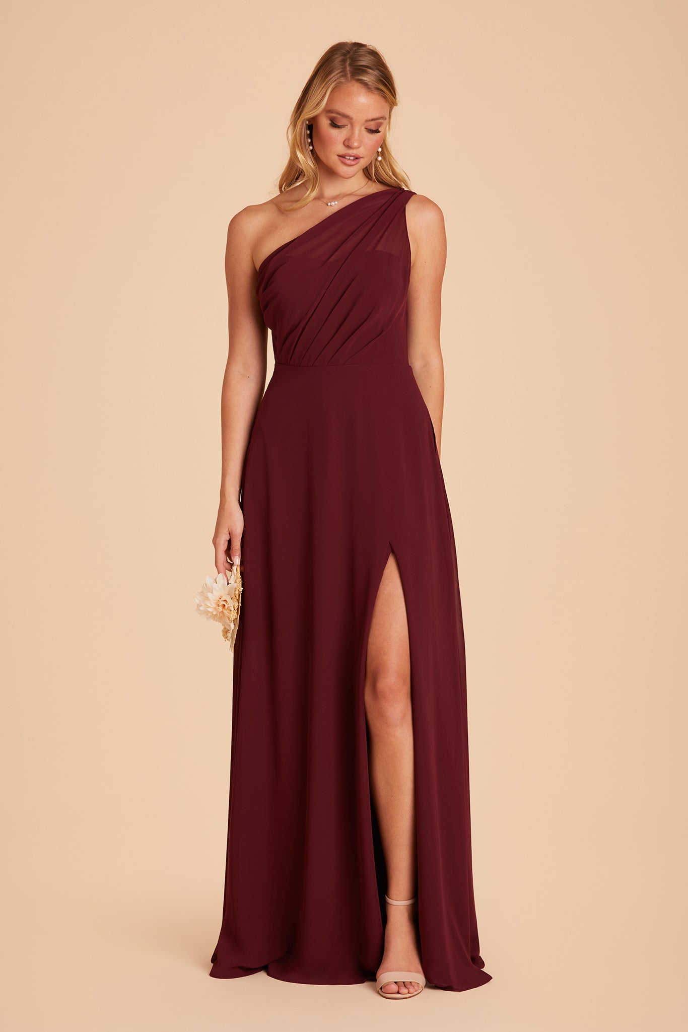 Front view of the Kira Dress in cabernet chiffon shows a slender model with a light skin tone wearing an asymmetrical one-shoulder, full-length dress with a mid-thigh slit on the left side. Chiffon gathers into soft pleating at the shoulder of the bodice with a smooth fit at the waist as the dress skirt with a slight A-line silhouette flows to the floor.    