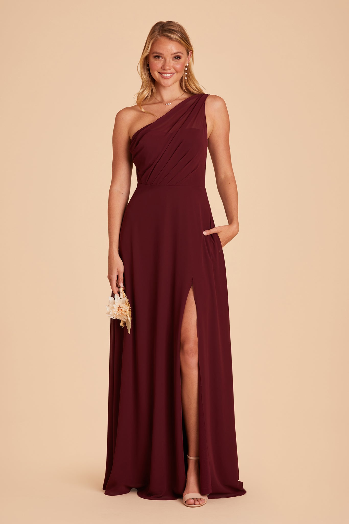 Front view of the Kira Dress in cabernet chiffon shows a slender model with a light skin tone with their hand tucked into their left pocket revealing their leg and sandled foot through the dress slit. The model wears the Mary High Chunky Heel shoe in nude blush.