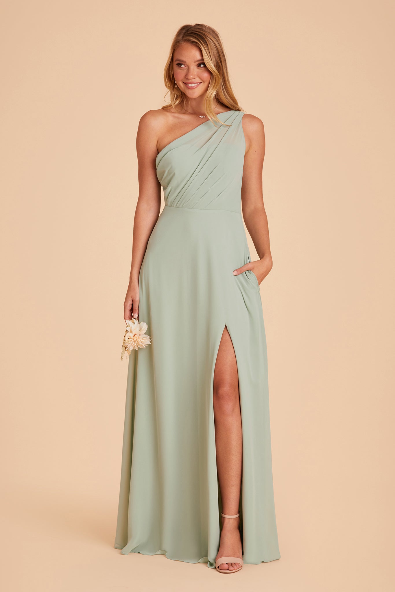 Front view of the Kira Dress in sage chiffon with the optional slit shows a slender model with a light skin tone wearing an asymmetrical one-shoulder, full-length dress. Soft pleating gathers at the left shoulder of the bodice with a smooth fit at the waist as the dress flows to the floor. 