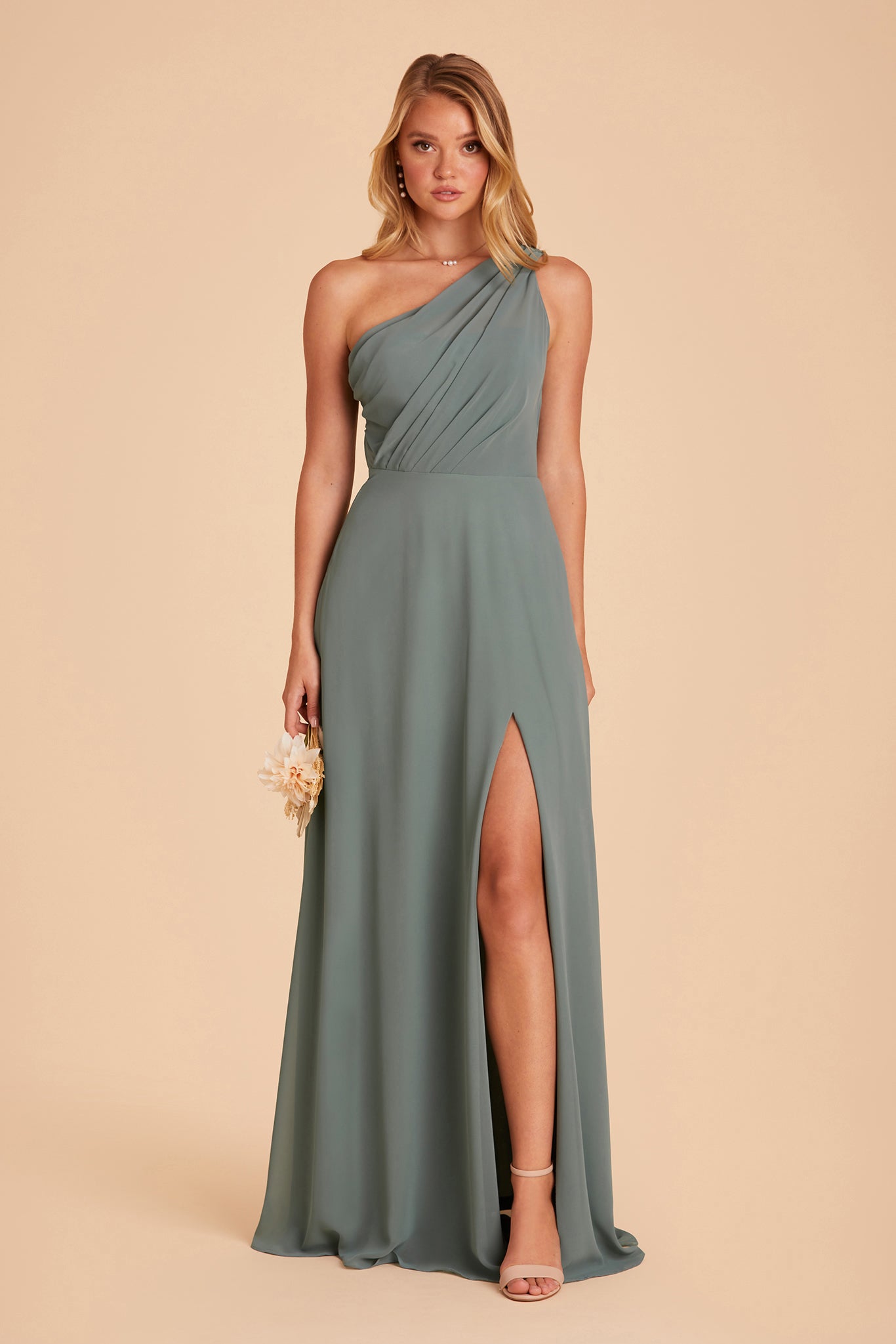 Front view of the Kira Dress in sea glass chiffon with the optional slit shows a slender model with a light skin tone with their left leg and foot revealed through a mid-thigh dress slit. They wear Natalie Chunky Heel shoes in nude blush. 