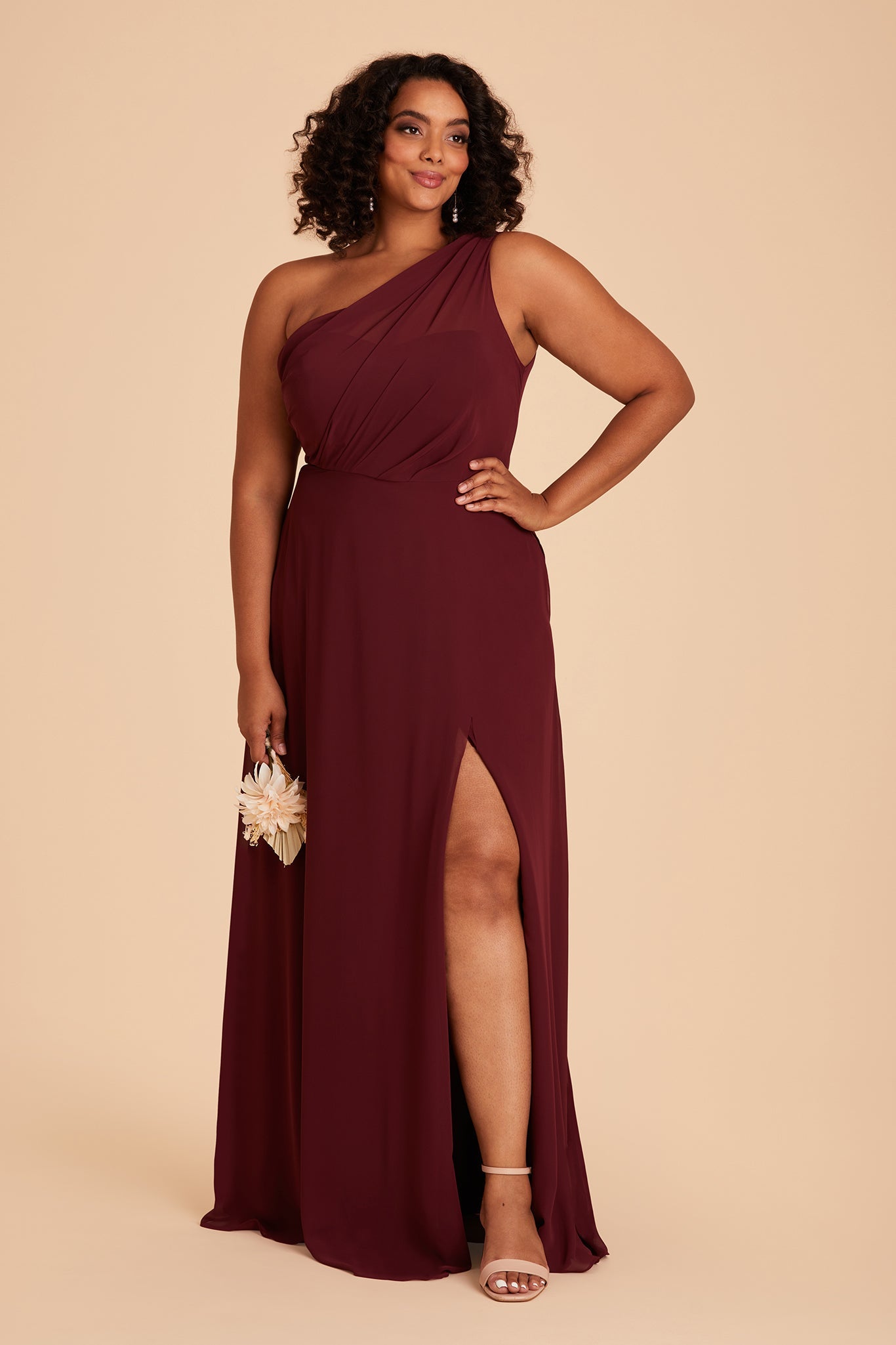 Front view of the Kira Dress Curve in cabernet chiffon with the optional slit shows a full-figured model with a medium skin tone wearing an asymmetrical one-shoulder, full-length dress. Soft pleating gathers at the left shoulder of the bodice with a smooth fit at the waist as the dress with a slight A-line silhouette flows to the floor.
