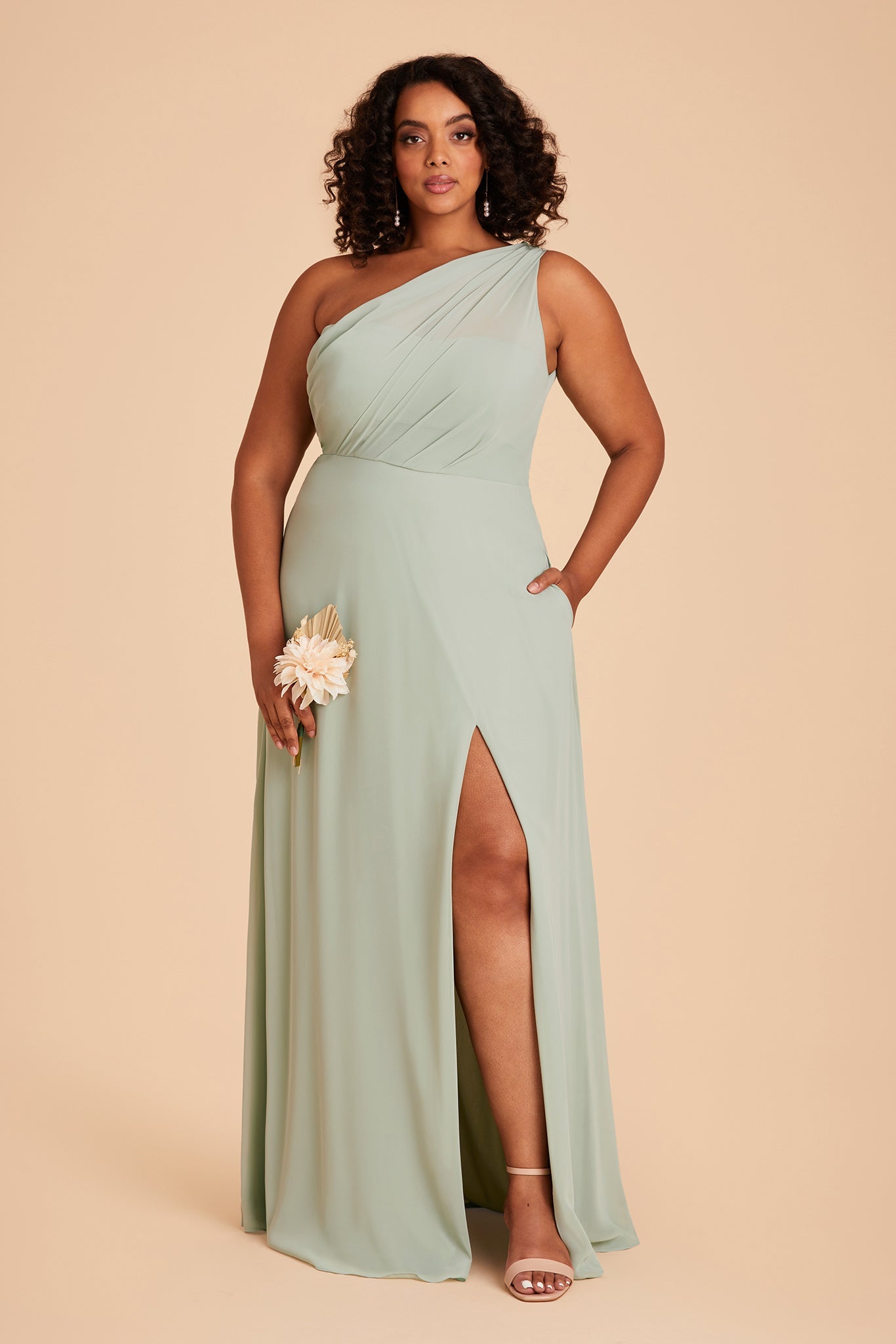 Front view of the Kira Dress Curve in sage chiffon with the optional slit shows a full-figured model with a medium skin tone wearing an asymmetrical one-shoulder, full-length dress. Soft pleating gathers at the left shoulder of the bodice with a smooth fit at the waist as the dress skirt flows to the floor.