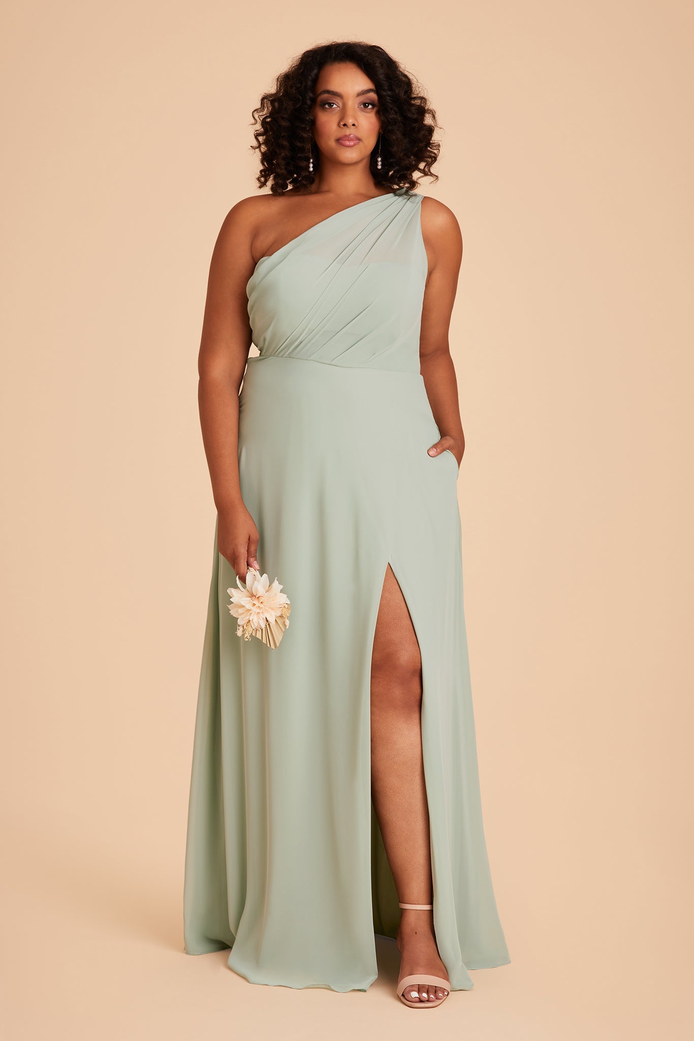 Front view of the Kira Dress Curve in sage chiffon with a slit shows the model with their left leg and foot revealed through a mid-thigh dress slit. They wear Natalie Chunky Heel shoes in nude blush.