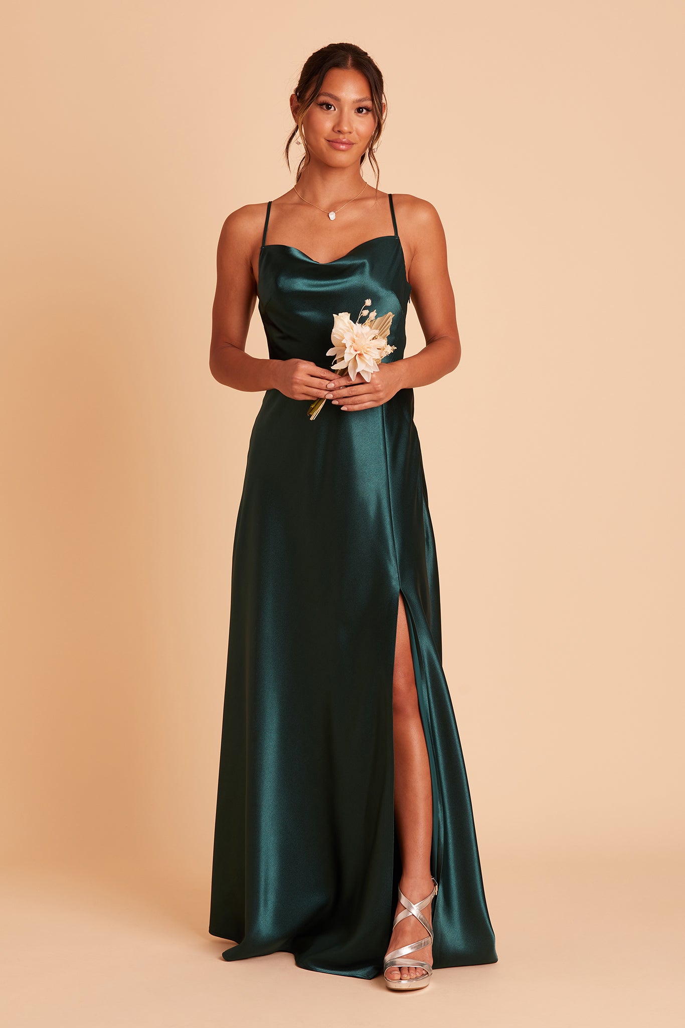 Front view of the Lisa Long Dress in emerald satin shows a model revealing their leg and foot in the mid-thigh slit. They wear the strappy high heel shoes in gold metallic.